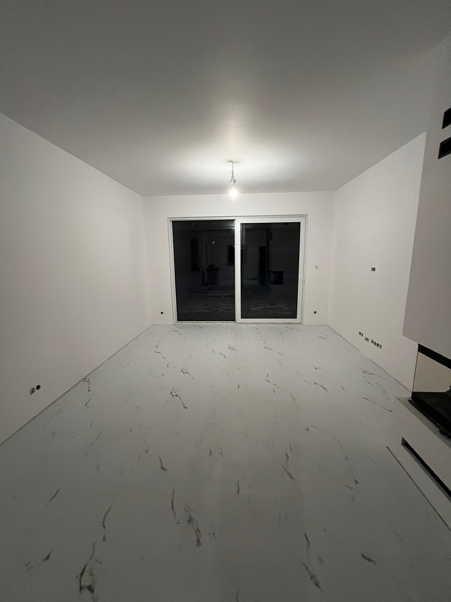 Wnętrze pokoju po zakończonych pracach, z białymi ścianami, marmurową podłogą w odcieniach szarości i czerni, oraz dużym przesuwnym oknem balkonowym. Widoczne gniazdka elektryczne na ścianie...