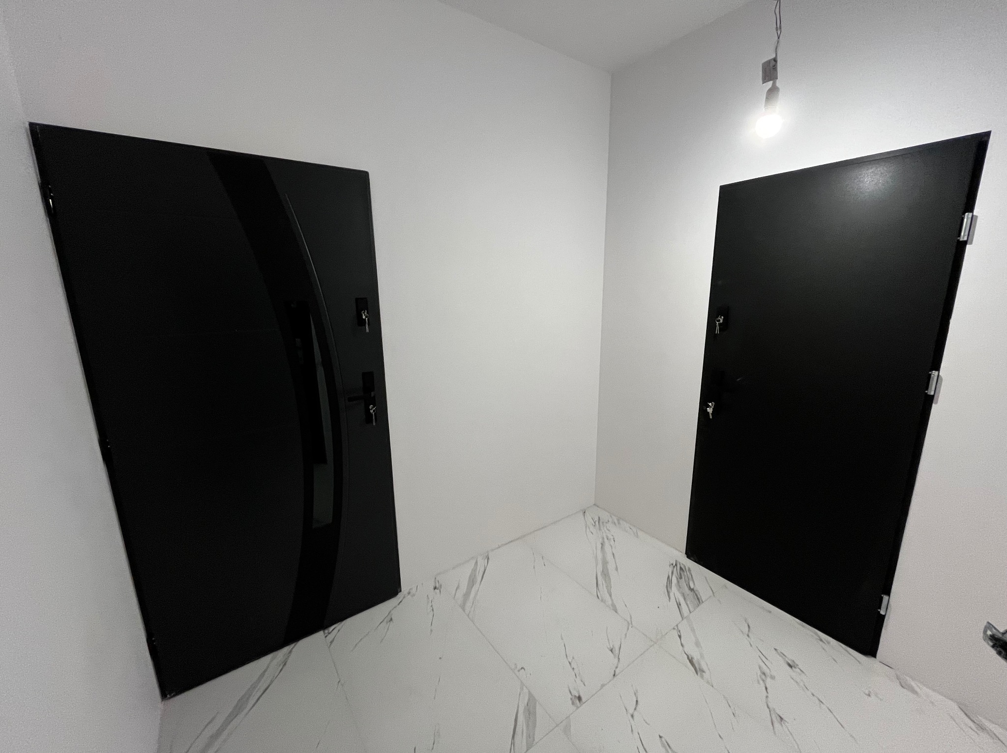 Dwa czarne, nowoczesne drzwi wewnętrzne w białym korytarzu z marmurową podłogą, jedno z przeszkleniem, oświetlone wiszącą żarówką.