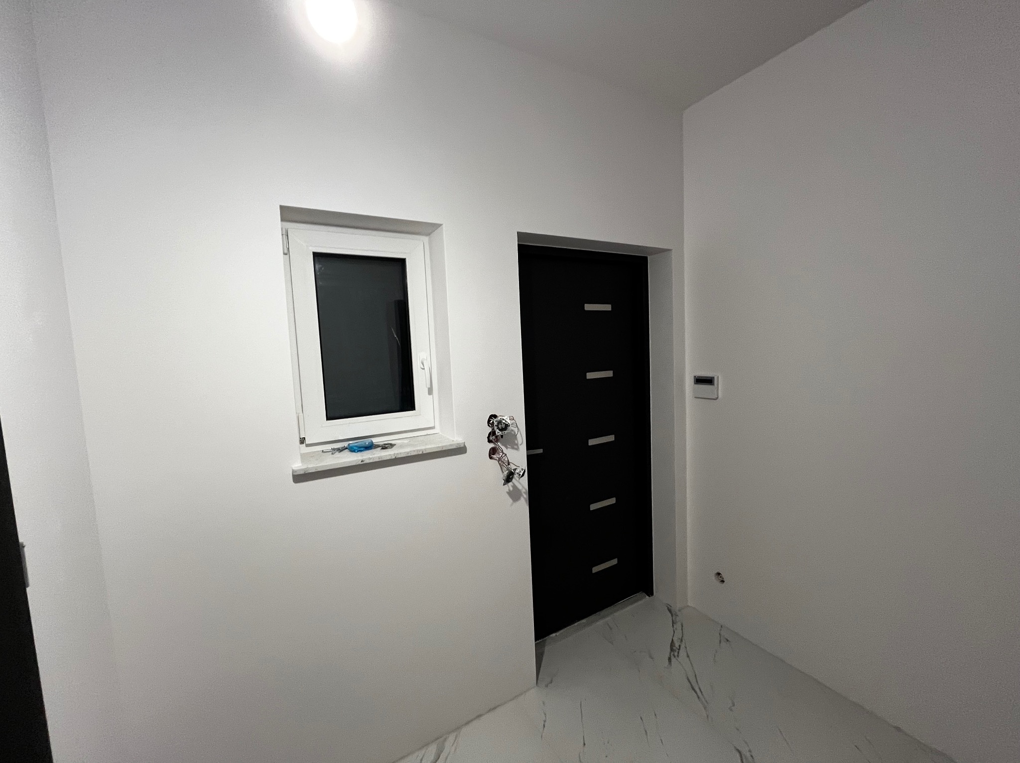 Wnętrze po świeżym malowaniu, widoczne białe ściany, czarne drzwi z poziomymi wgłębieniami, białe okno z narzędziem na parapecie oraz marmurowe płytki podłogowe.