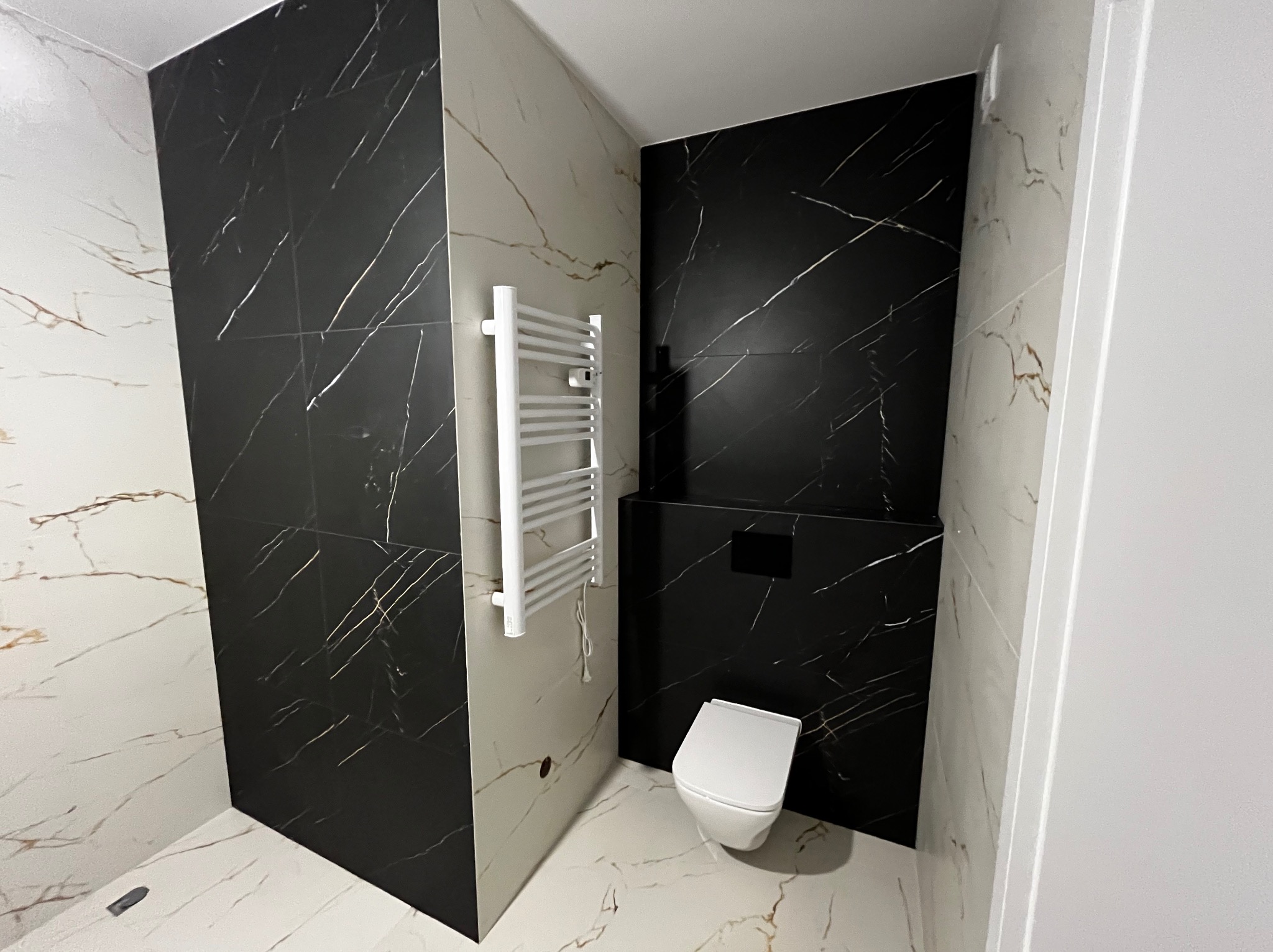 Wykończona łazienka z czarno-białymi płytkami imitującymi marmur, wiszącym grzejnikiem drabinkowym i podwieszaną toaletą.
