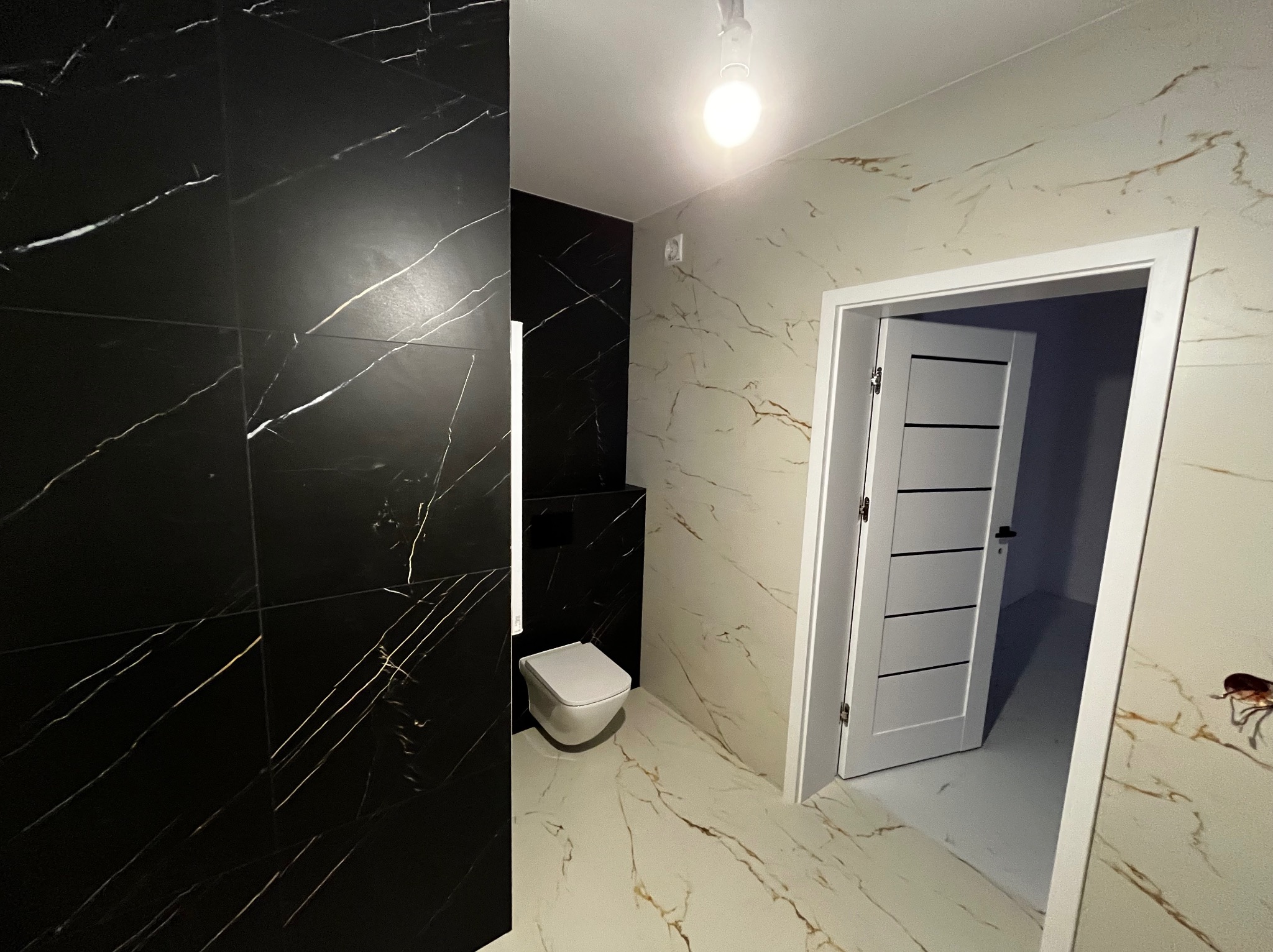 Nowoczesna łazienka z czarnymi płytkami imitującymi marmur na ścianie, wisząca toaleta i drzwi z poziomymi frezami, wykończona w beżowo-złotej kolorystyce.