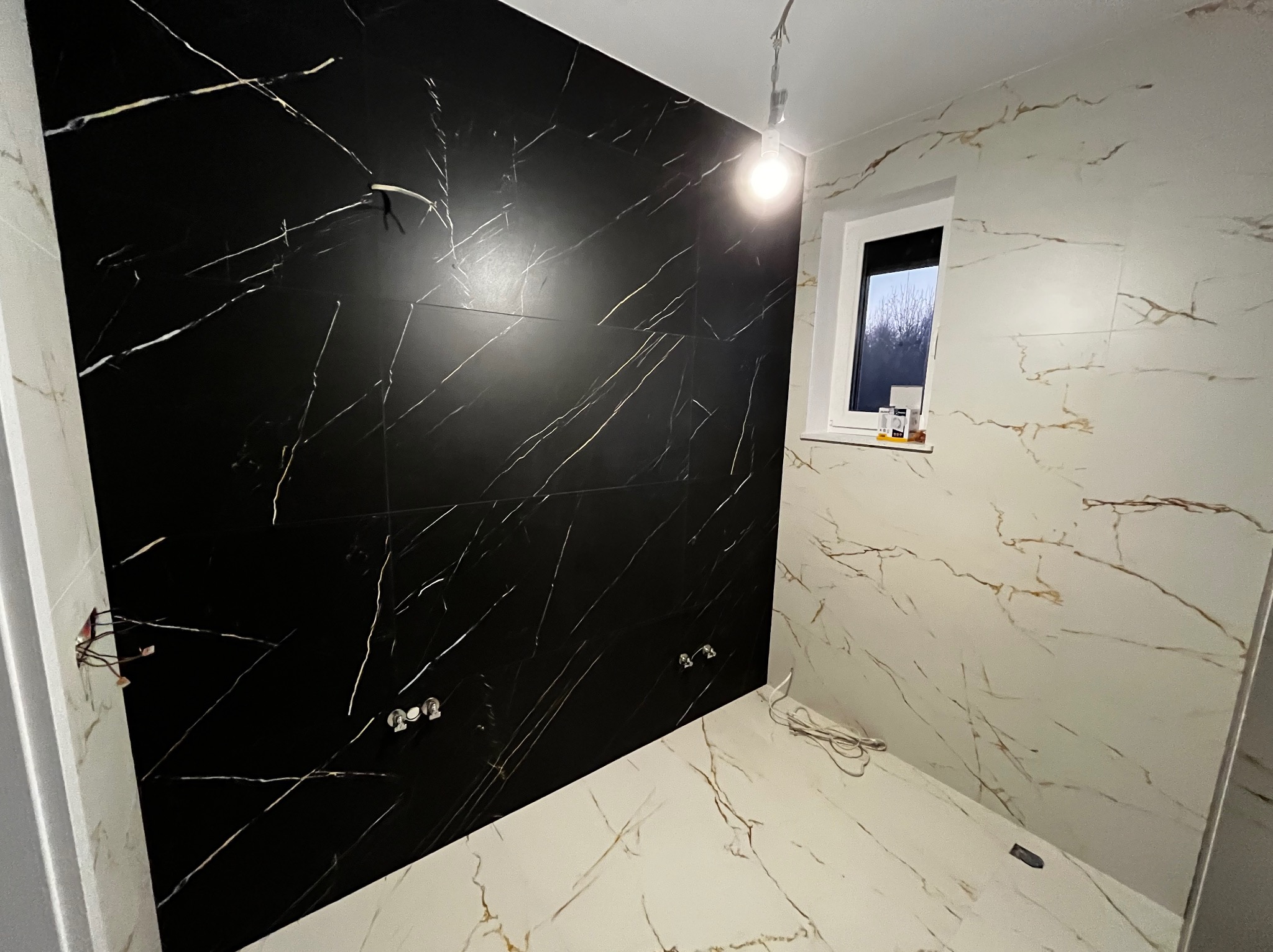 Łazienka w trakcie wykańczania, z czarną ścianą z płytek imitujących marmur z białymi żyłami, białe płytki na pozostałych ścianach i podłodze również imitujące marmur, widoczne wystające kable...