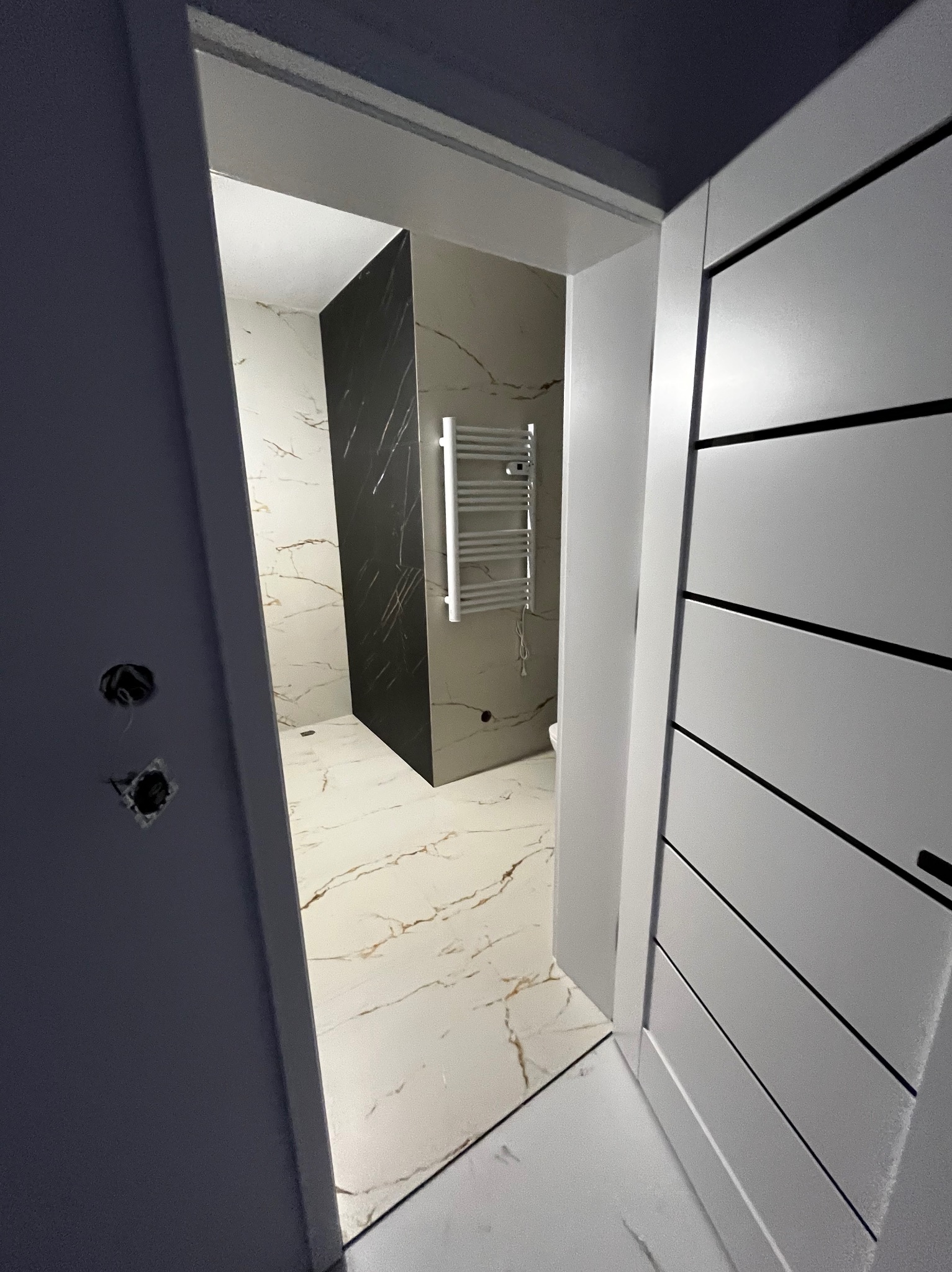 Widok na wykończoną łazienkę przez otwarte drzwi z nowoczesnym grzejnikiem drabinkowym, marmurową okładziną ścian i podłogi, oraz widocznymi puszkami elektrycznymi na ścianie obok ościeżnicy.