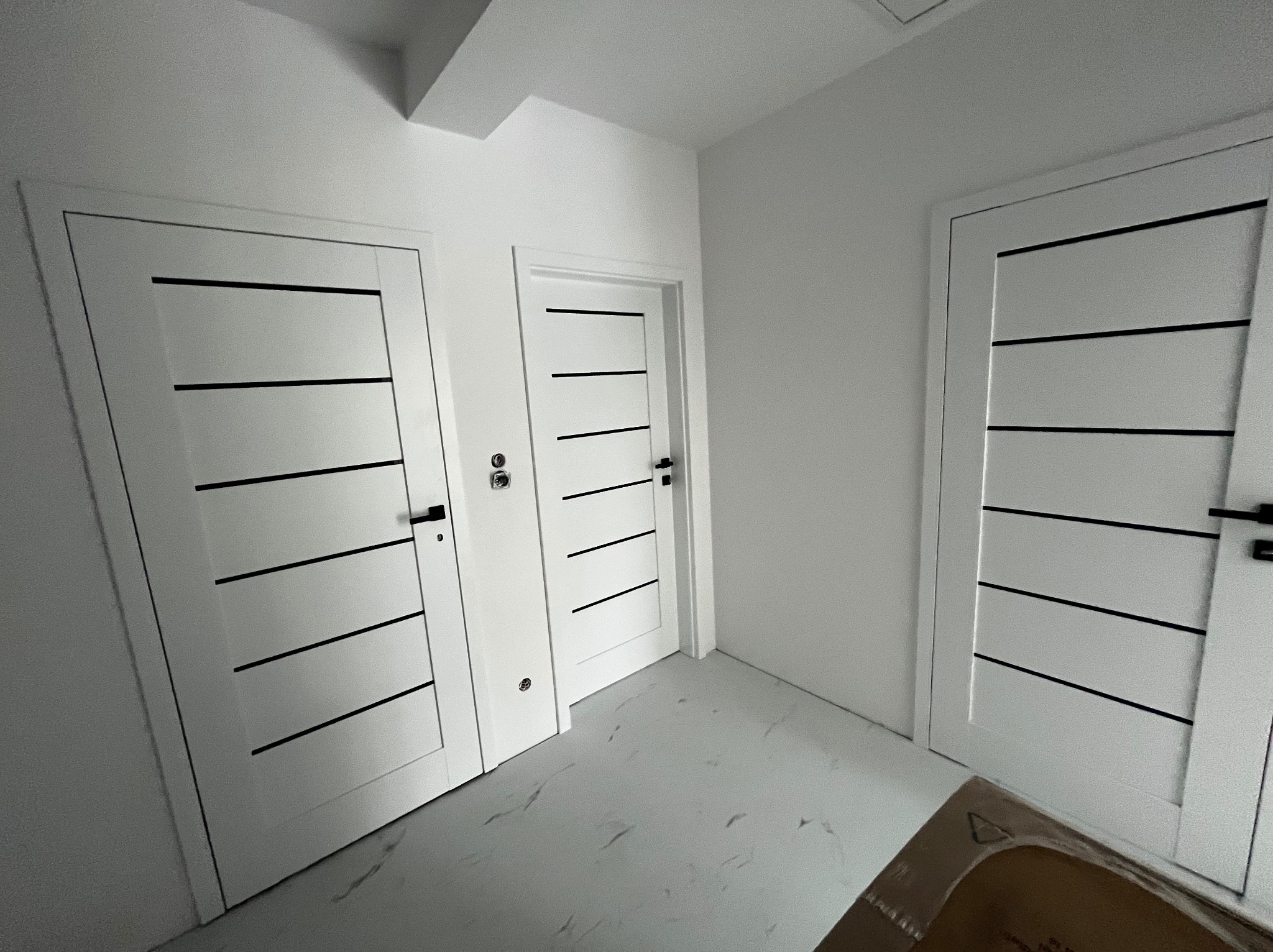 Trzy białe, nowoczesne drzwi wewnętrzne z czarnymi, poziomymi paskami w nowo wyremontowanym, jasnym pomieszczeniu z podłogą imitującą marmur.