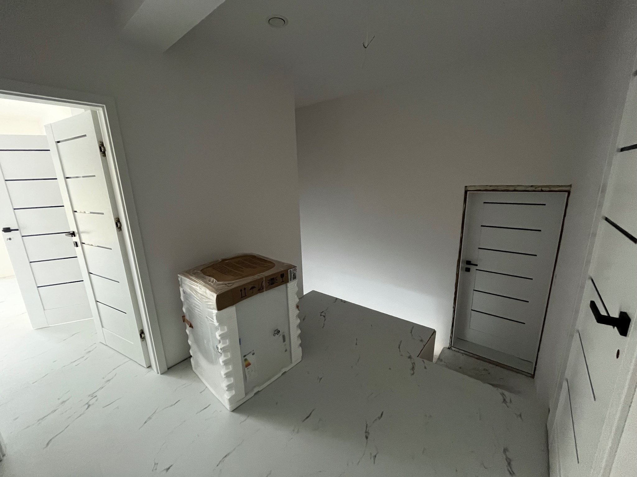 Wnętrze w trakcie wykańczania: białe ściany, marmurowe płytki podłogowe, drzwi z czarnymi poziomymi paskami, zamontowane ościeżnice i zapakowana w folię umywalka z szafką.