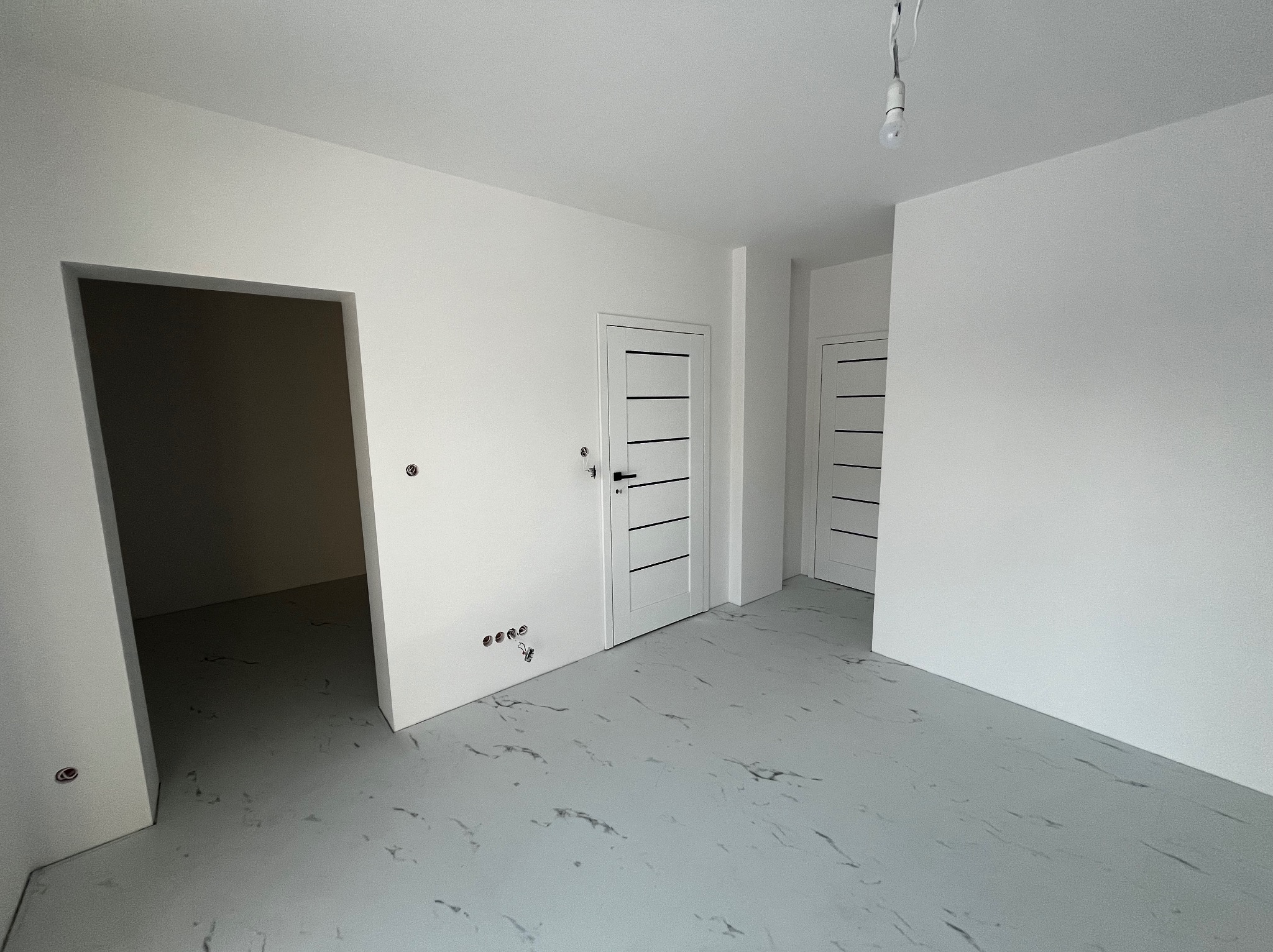 Wnętrze po remoncie: białe ściany, szara podłoga imitująca marmur, dwoje białych drzwi z czarnymi poziomymi paskami, widoczne gniazdka elektryczne i wisząca żarówka.