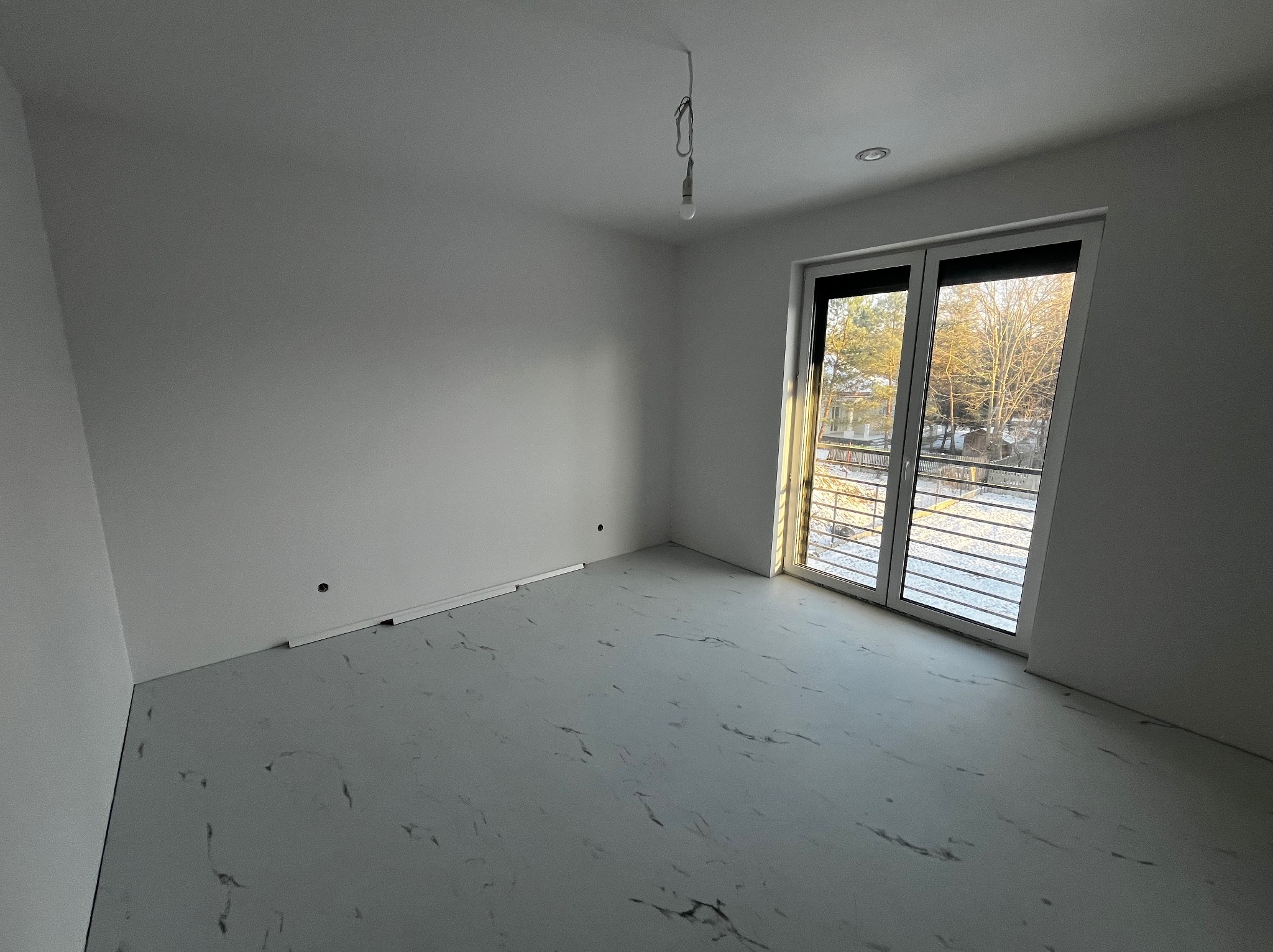 Pusty, odnowiony pokój z białymi ścianami, szarą podłogą imitującą marmur i dużymi, przeszklonymi drzwiami balkonowymi z widokiem na zewnątrz, z wiszącą żarówką na kablu.