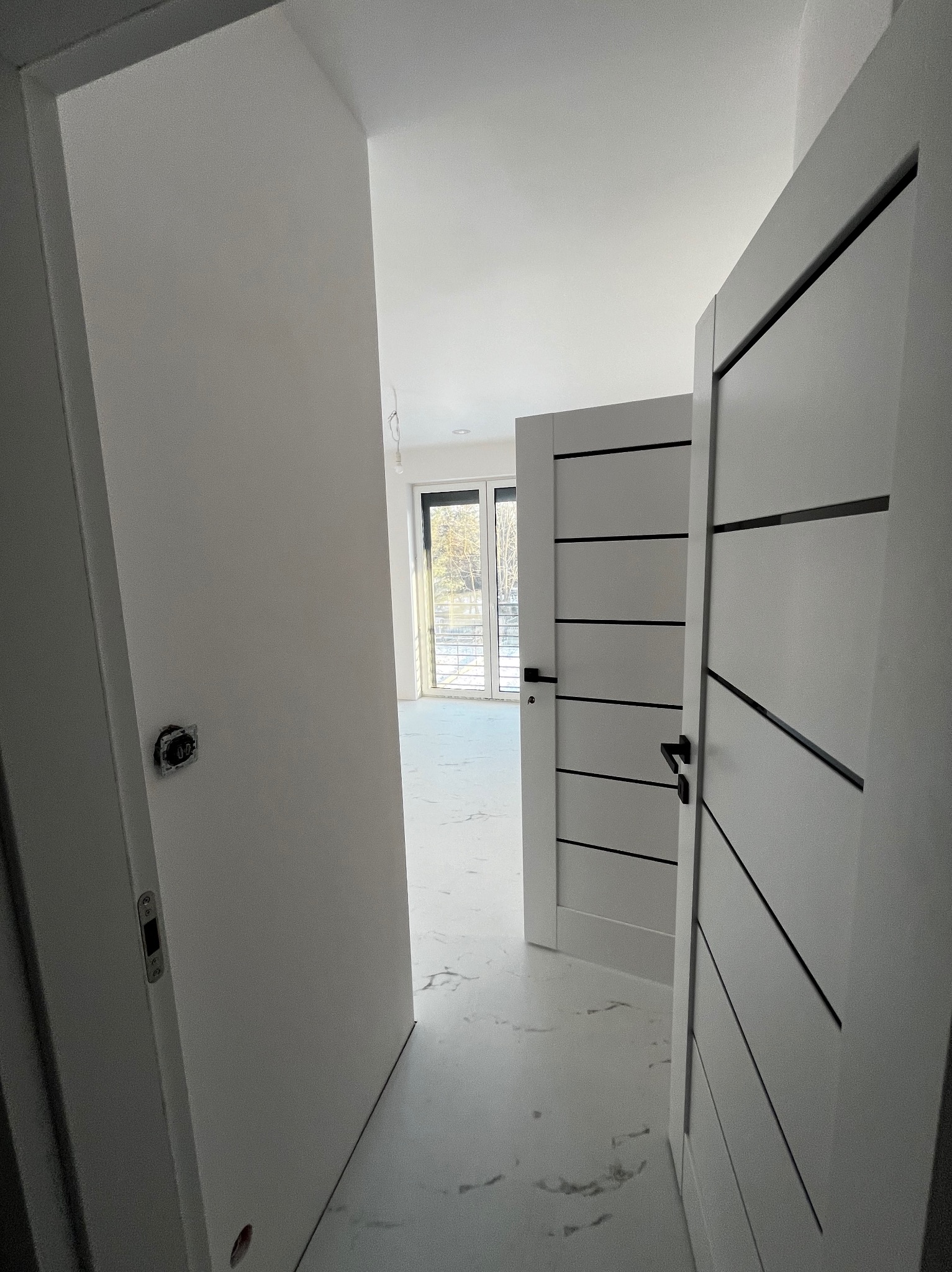 Wnętrze w trakcie prac wykończeniowych; widoczne białe ściany, marmurowa podłoga zabezpieczona folią, oraz nowoczesne, szare drzwi z czarnymi wstawkami i klamkami.