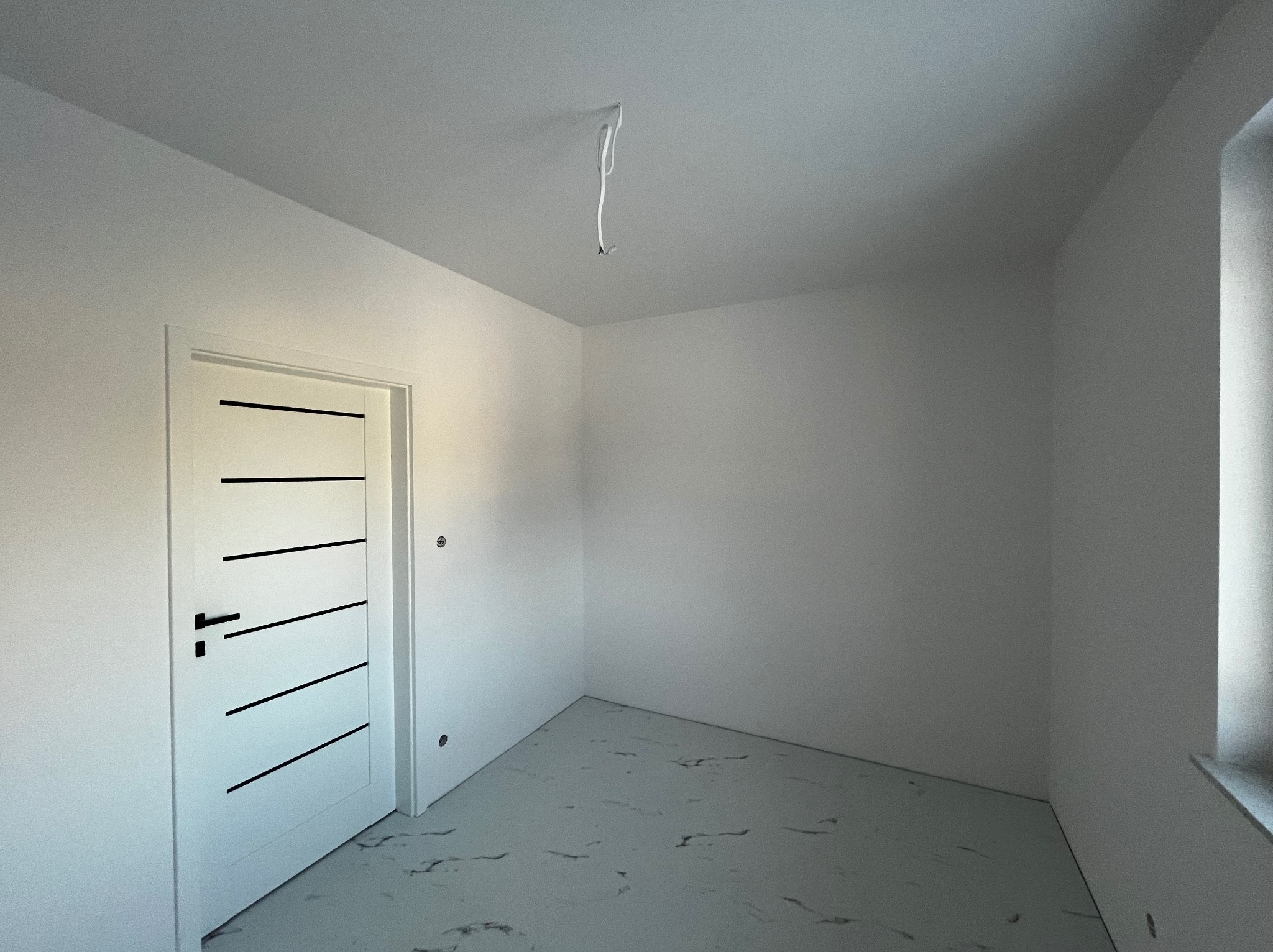 Pusty, biały pokój z nowymi drzwiami z czarnymi poziomymi paskami, marmurową podłogą i wystającymi kablami z sufitu.