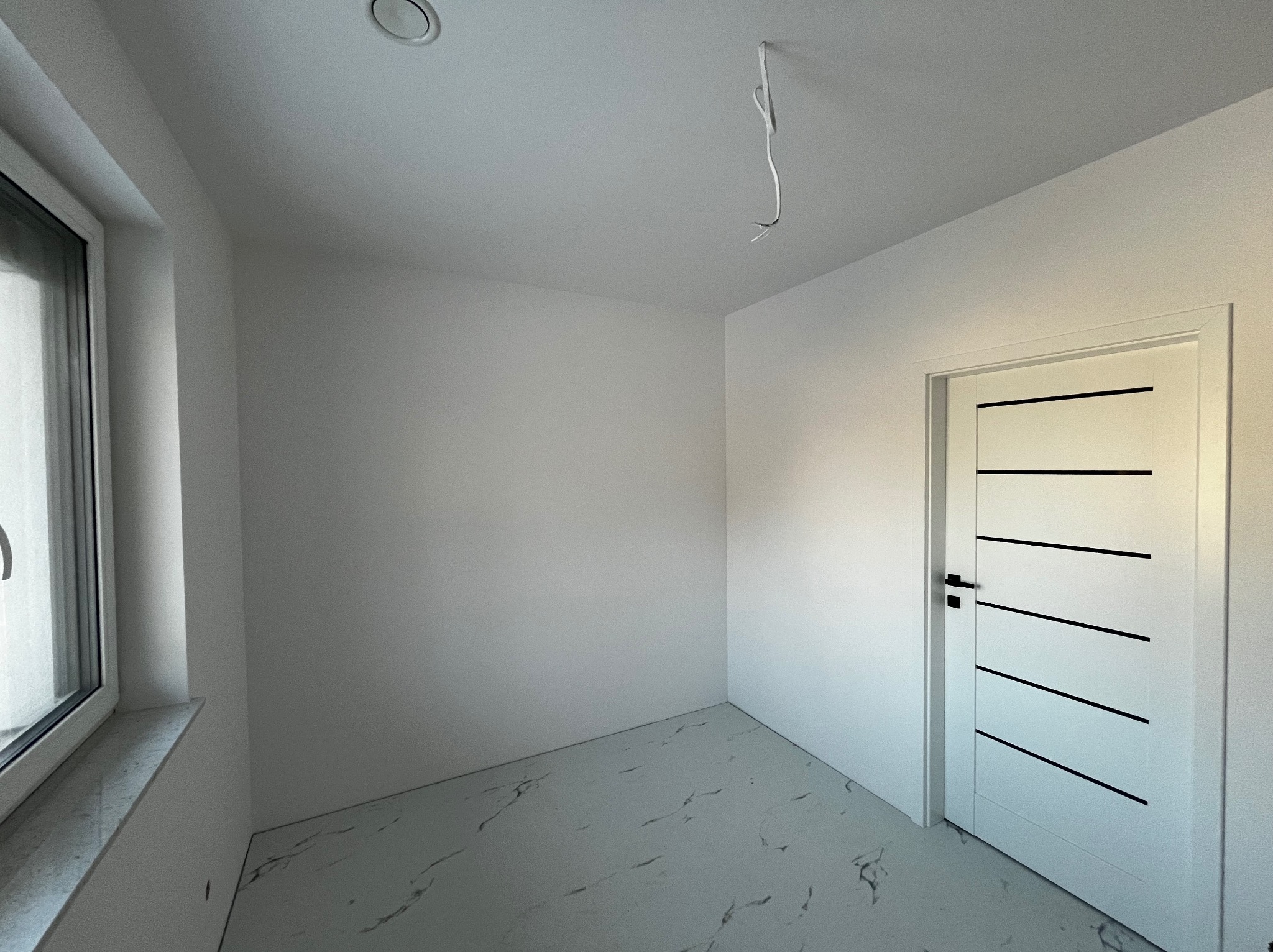 Pomalowane na biało wnętrze pokoju z białymi ścianami, marmurową podłogą, oknem i nowoczesnymi białymi drzwiami z czarnymi poziomymi paskami, z wystającym kablem z sufitu.