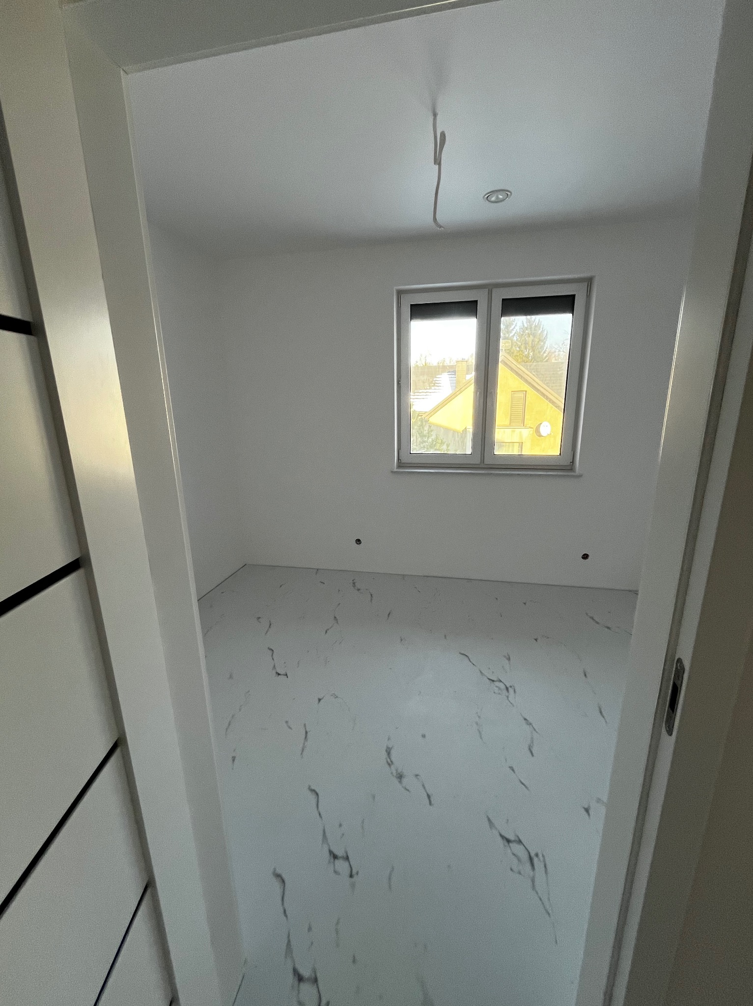 Pusty pokój z białymi ścianami i podłogą imitującą marmur, widok przez otwarte drzwi z czarnymi akcentami, okno z widokiem na żółty dom na zewnątrz, wystający kabel z sufitu.