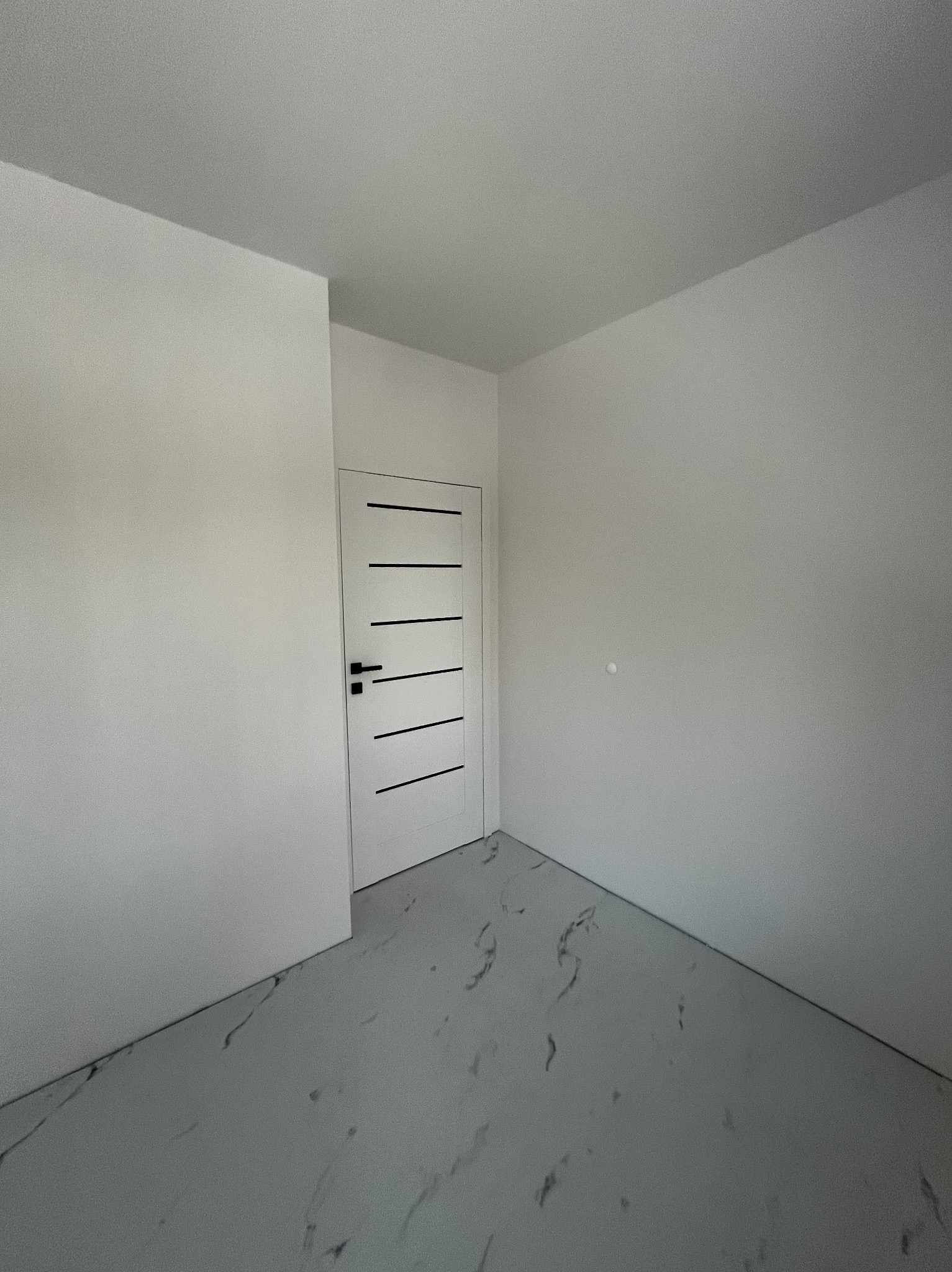 Wykończone wnętrze pokoju z białymi ścianami, szarą podłogą imitującą marmur i zamontowanymi białymi drzwiami z czarnymi, poziomymi paskami.