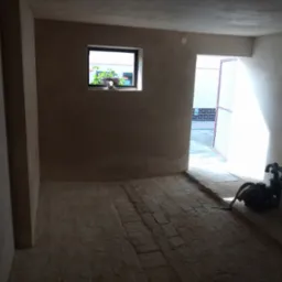 Wnętrze pomieszczenia po nałożeniu tynku, widoczna faktura ściany, okno z kwiatami i sprzęt do tynkowania w rogu.