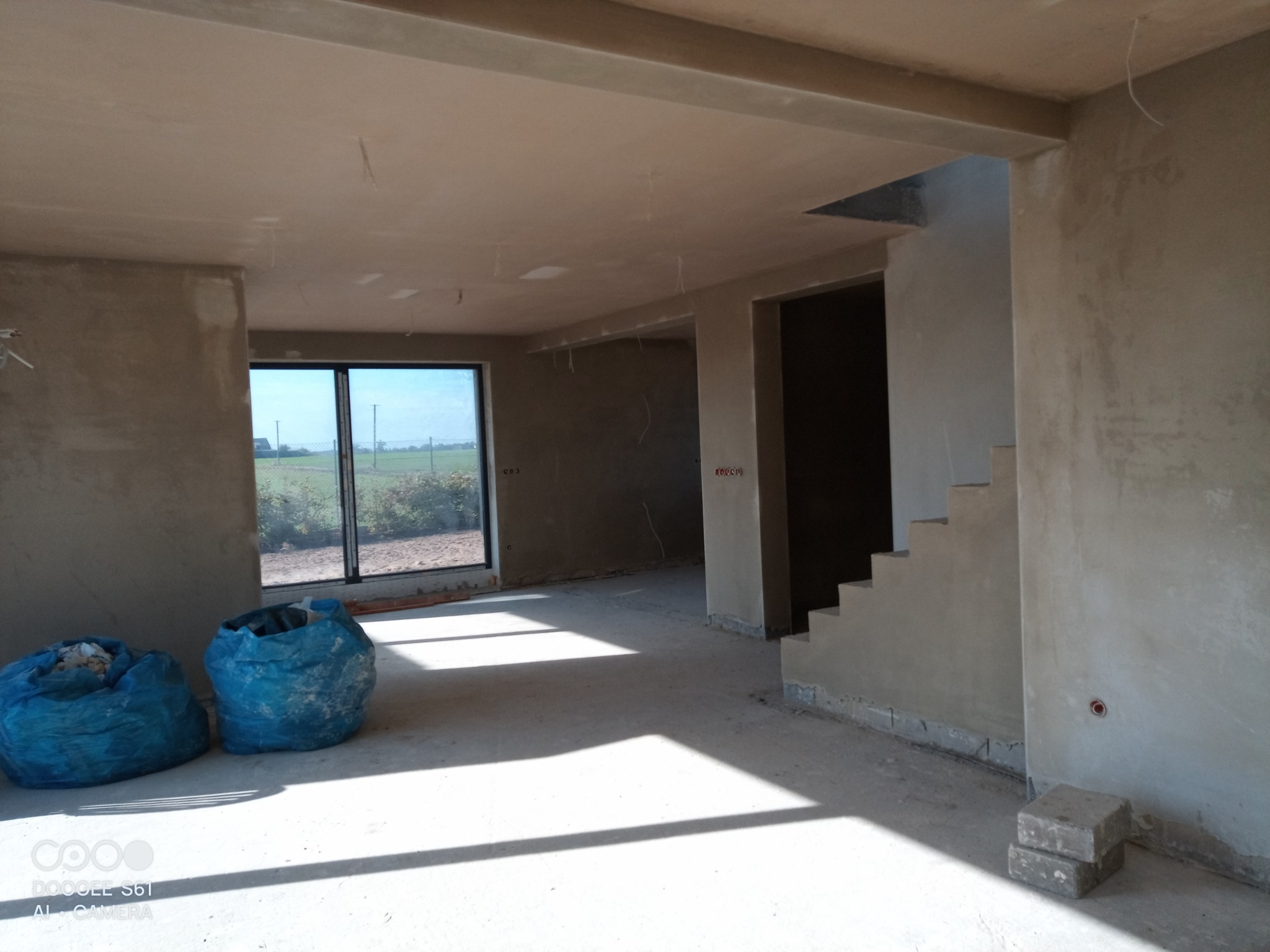 Wnętrze domu w trakcie prac wykończeniowych, ściany i sufit pokryte świeżym tynkiem, widoczne dwa worki z resztkami materiałów budowlanych oraz fragment schodów.