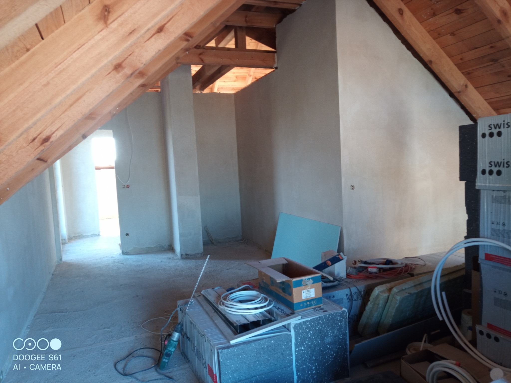 Poddasze w trakcie tynkowania maszynowego, widoczne drewniane elementy konstrukcyjne dachu, ściany pokryte świeżym tynkiem, materiały budowlane i narzędzia na podłodze.