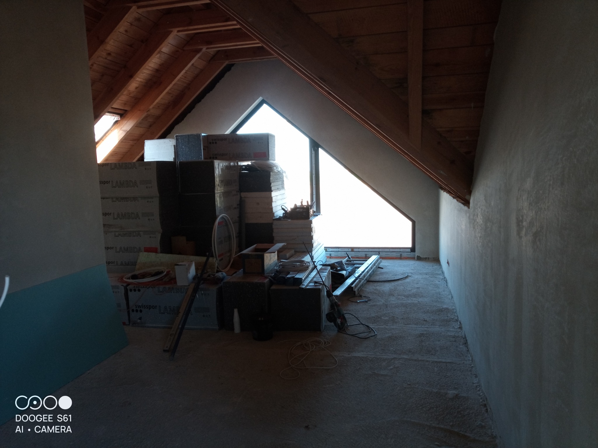 Poddasze w trakcie prac wykończeniowych, widoczne ocieplone skosy, okno dachowe, materiały budowlane i świeżo nałożony tynk na ścianie szczytowej.