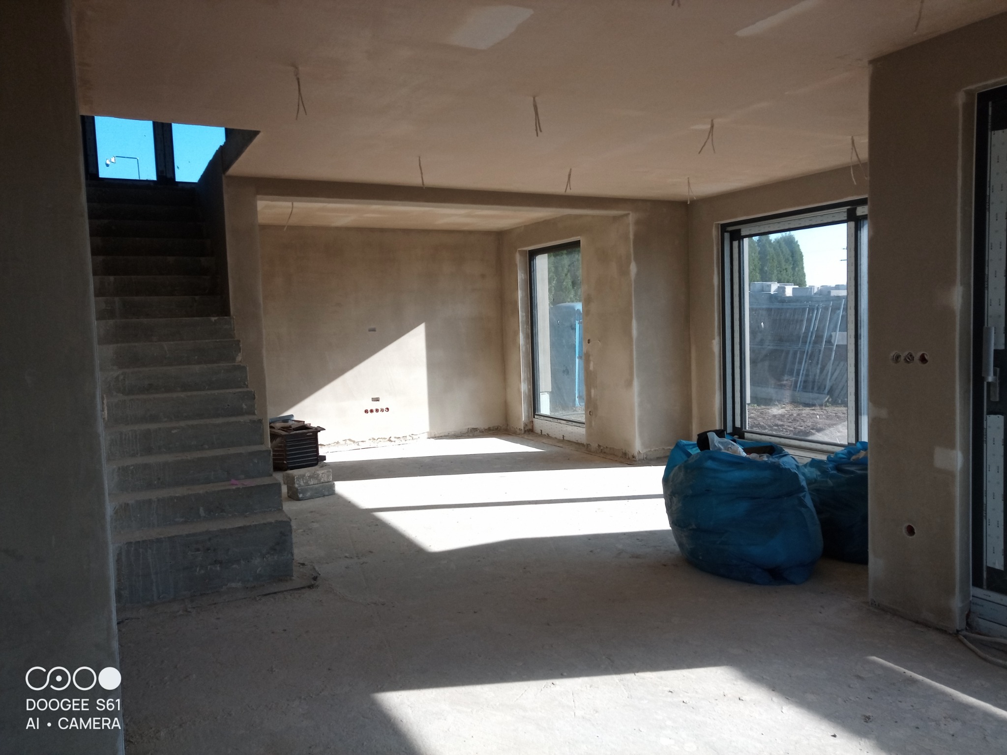 Wnętrze surowego domu po nałożeniu tynków, widoczne schody betonowe, okna z widokiem na zewnątrz i worki z odpadami budowlanymi na podłodze.