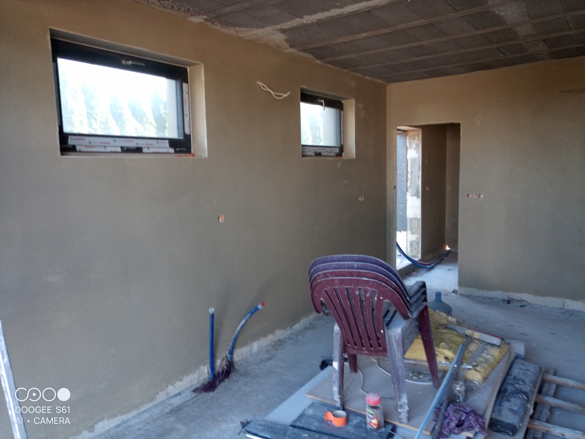 Wnętrze domu z otynkowanymi ścianami, widoczne dwa okna z czarnymi ramami, betonowy sufit, sterta krzeseł i materiały budowlane na podłodze, rury instalacji wodnej przy ścianie.