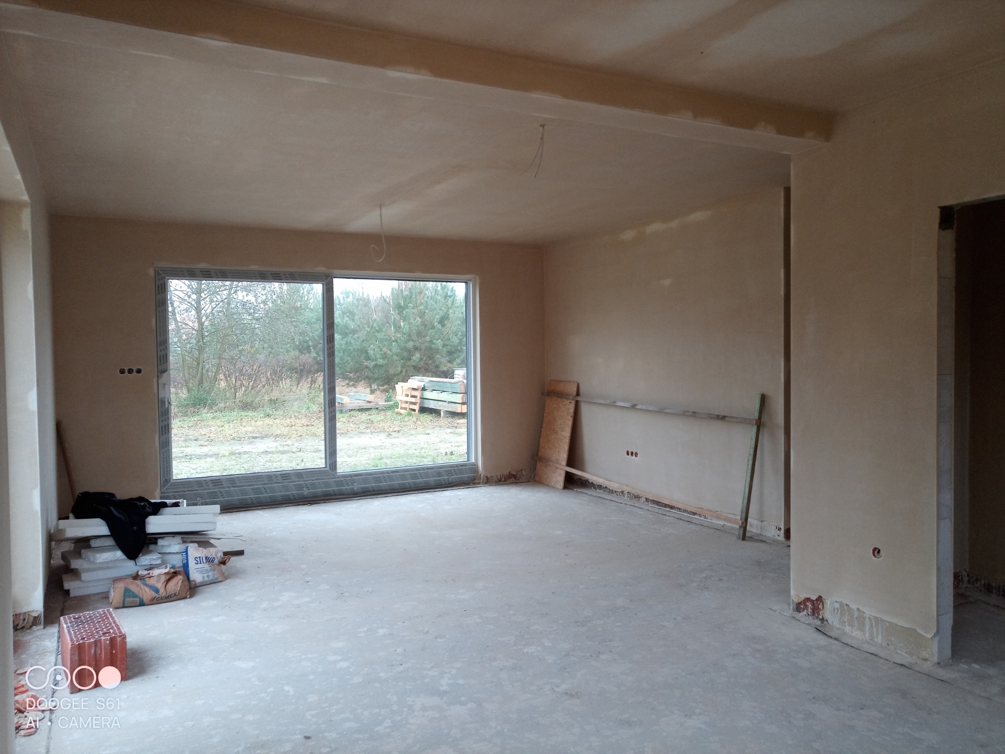 Wnętrze surowego domu po nałożeniu gładzi gipsowej, widoczne worki z cementem i bloczki na podłodze, duże okno z widokiem na zewnątrz.