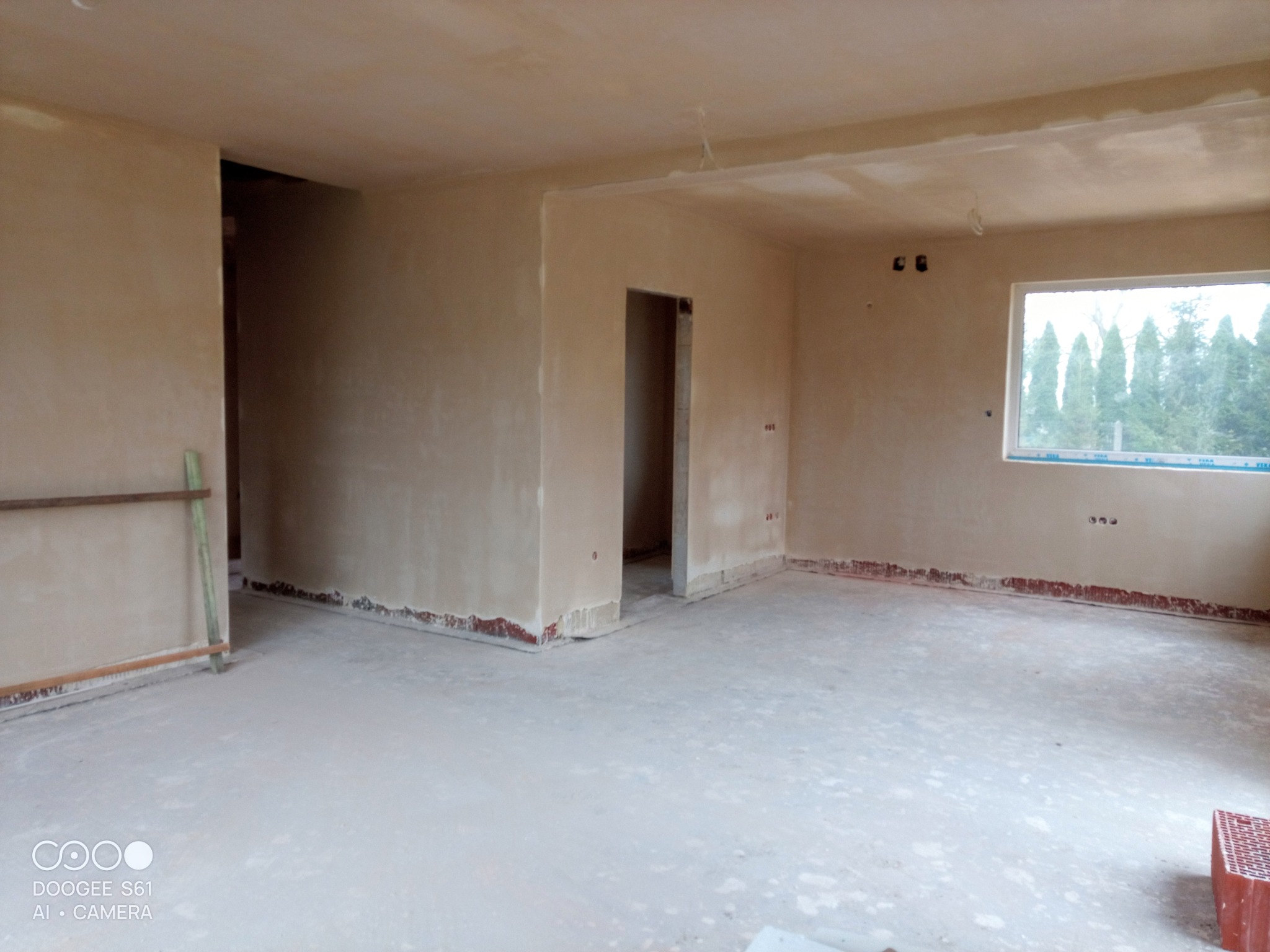 Wnętrze domu po nałożeniu tynków gipsowych, widoczne ślady zabezpieczeń przy podłodze, surowe wykończenie, okno z widokiem na zieleń.