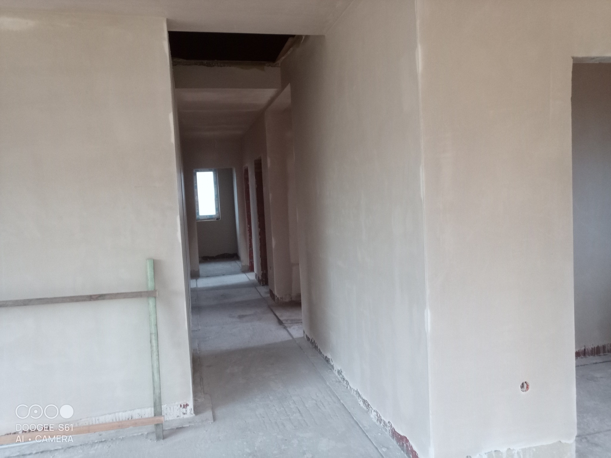 Wnętrze budynku w trakcie prac wykończeniowych: świeżo nałożone, jasne tynki na ścianach korytarza, widoczne drzwi do pomieszczeń, surowy stan podłogi i fragment rusztowania.