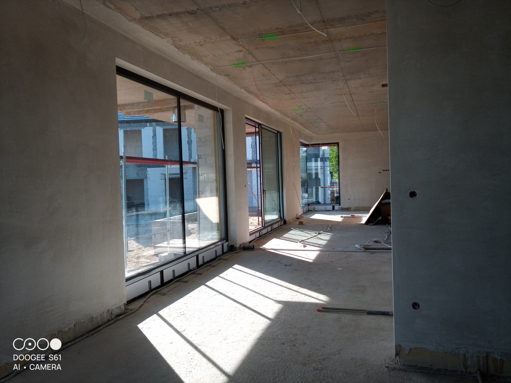 Wnętrze surowego budynku po nałożeniu tynków, z widocznymi dużymi oknami i promieniami słonecznymi padającymi na podłogę. Ściany pokryte szarym tynkiem, sufit betonowy z wystającymi przewodami...