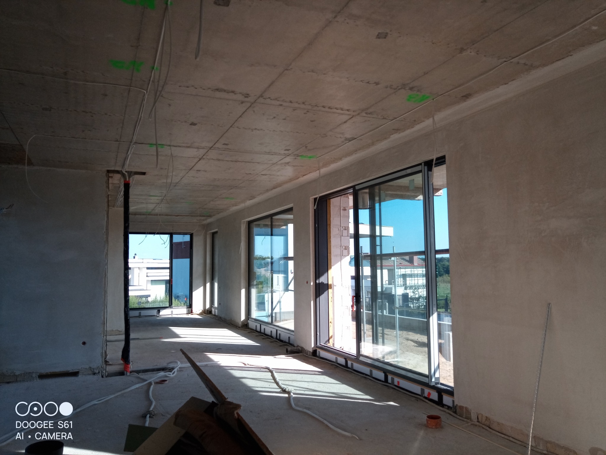 Surowe wnętrze w trakcie wykańczania, ściany pokryte świeżym tynkiem, duże okna przesuwne, widoczne przewody elektryczne pod sufitem, podpora stropu w lewym rogu.