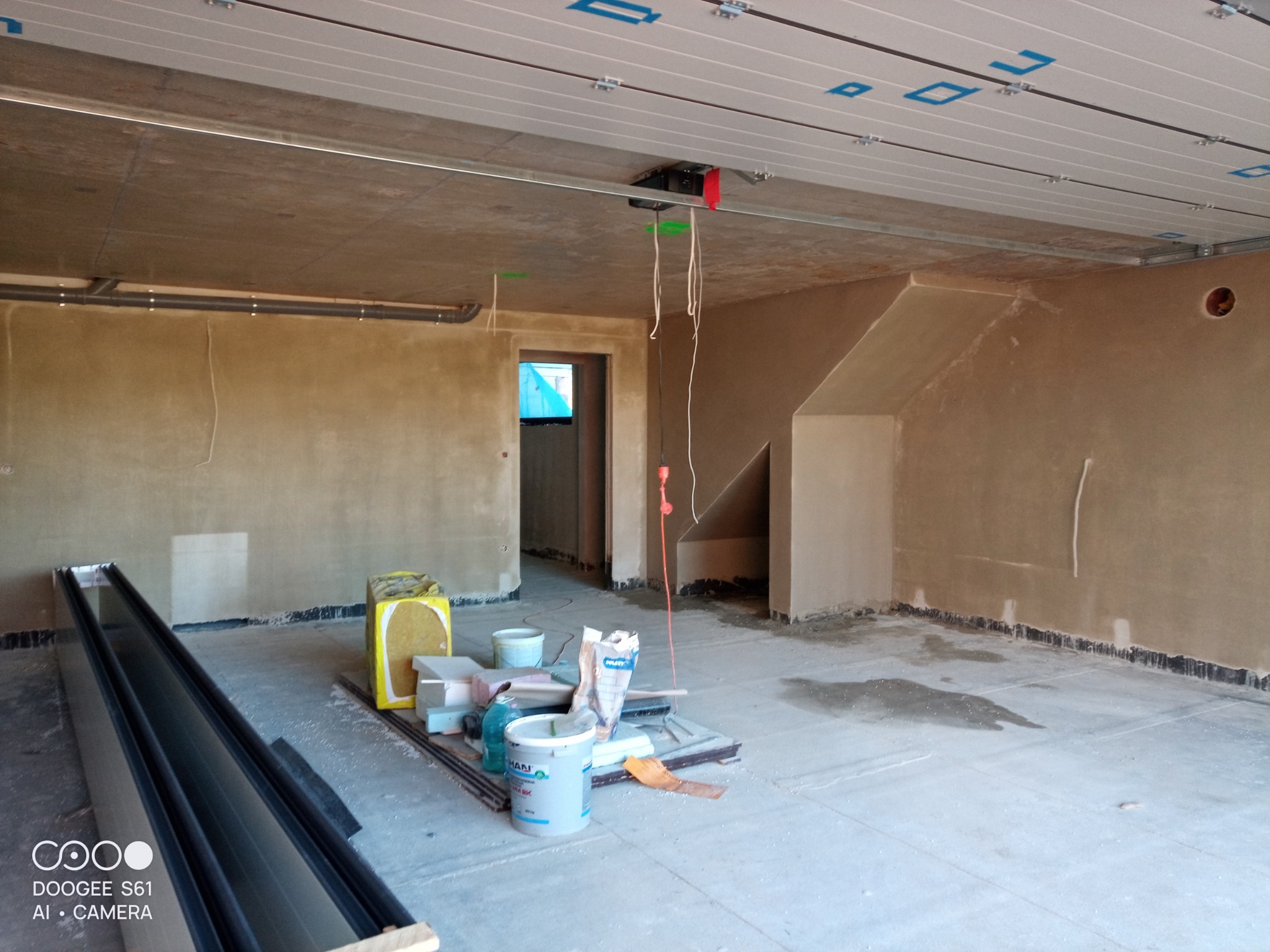 Wnętrze budynku w trakcie prac tynkarskich maszynowych, widoczna ściana po nałożeniu tynku, instalacje pod sufitem, narzędzia i materiały przygotowane do dalszej pracy.