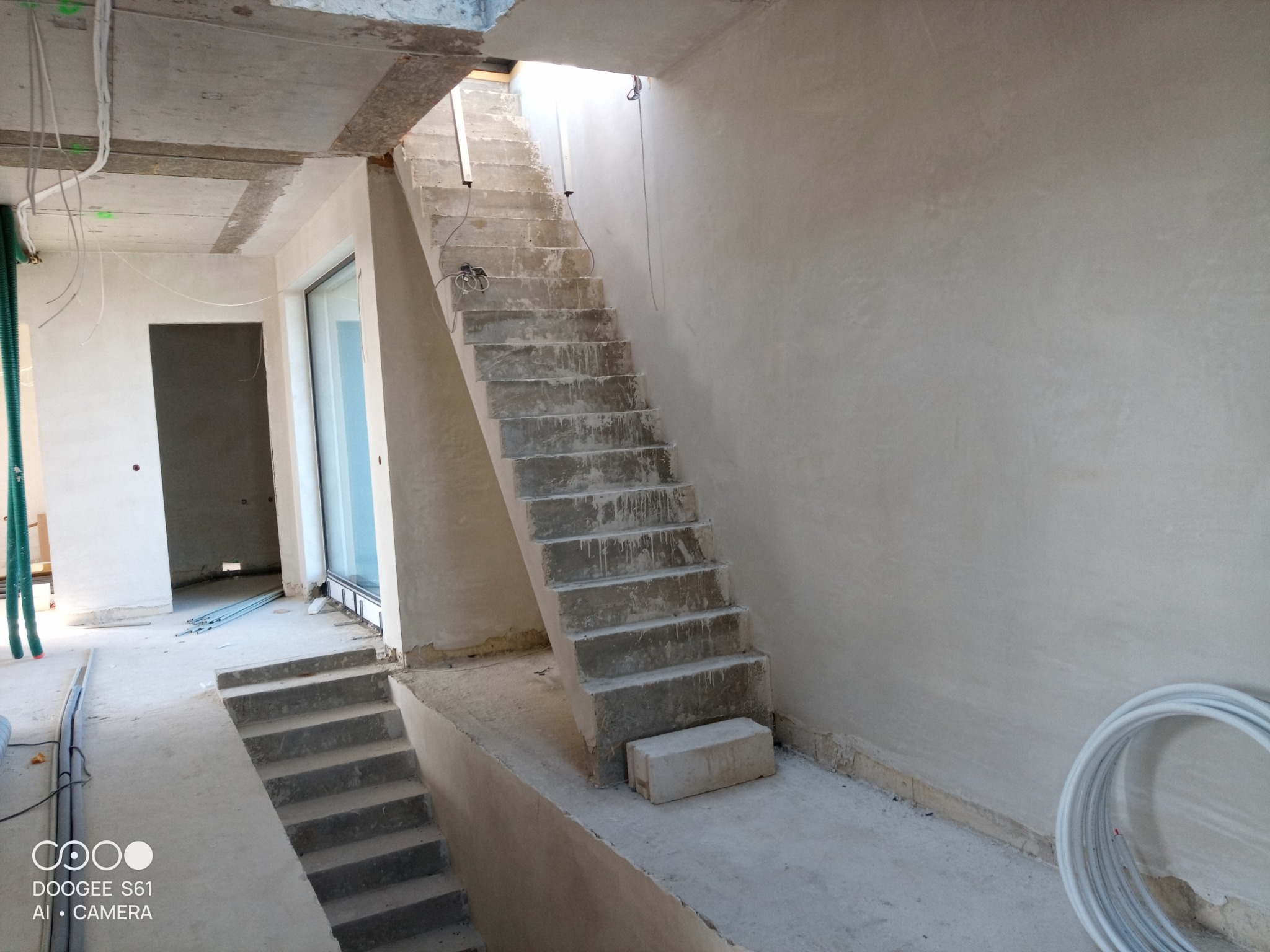 Wnętrze budynku w trakcie prac wykończeniowych, widoczne surowe schody betonowe, ściany pokryte świeżym tynkiem, oraz instalacje elektryczne i sanitarne.
