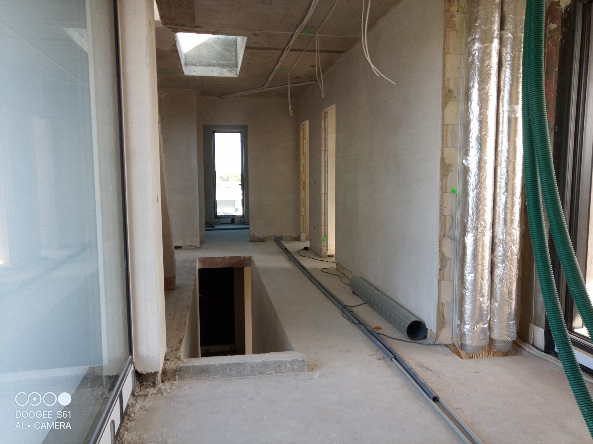 Surowe wnętrze w stanie deweloperskim po nałożeniu gładzi, widoczne przewody elektryczne pod sufitem, rury wentylacyjne w folii aluminiowej i otwór w podłodze.