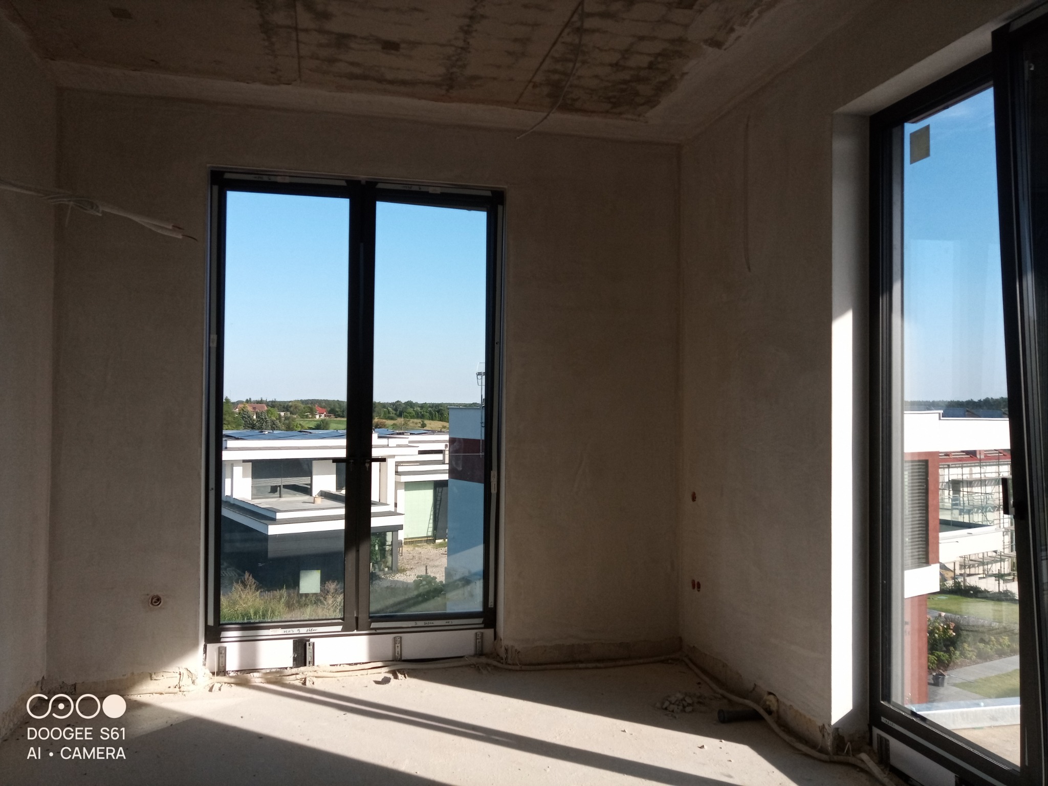 Wnętrze w stanie deweloperskim po nałożeniu tynków, widoczne ślady po instalacjach elektrycznych i rury przy podłodze, przez okno widok na nowoczesne budynki.