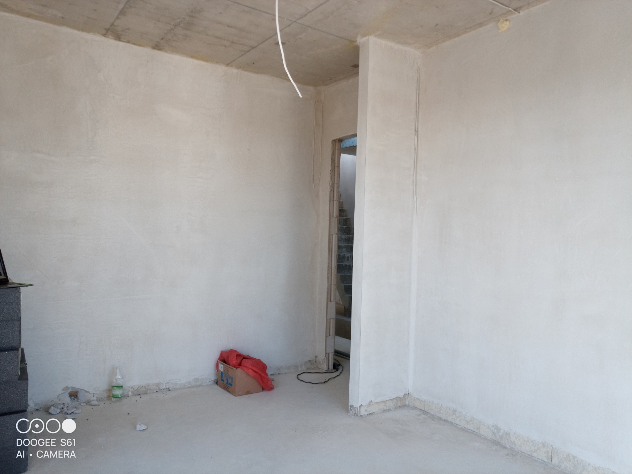 Surowe wnętrze po nałożeniu jasnoszarej warstwy tynku maszynowego, widoczna surowa betonowa podłoga i sufit, wystające przewody elektryczne, kartonowe pudło z czerwonymi elementami w rogu...