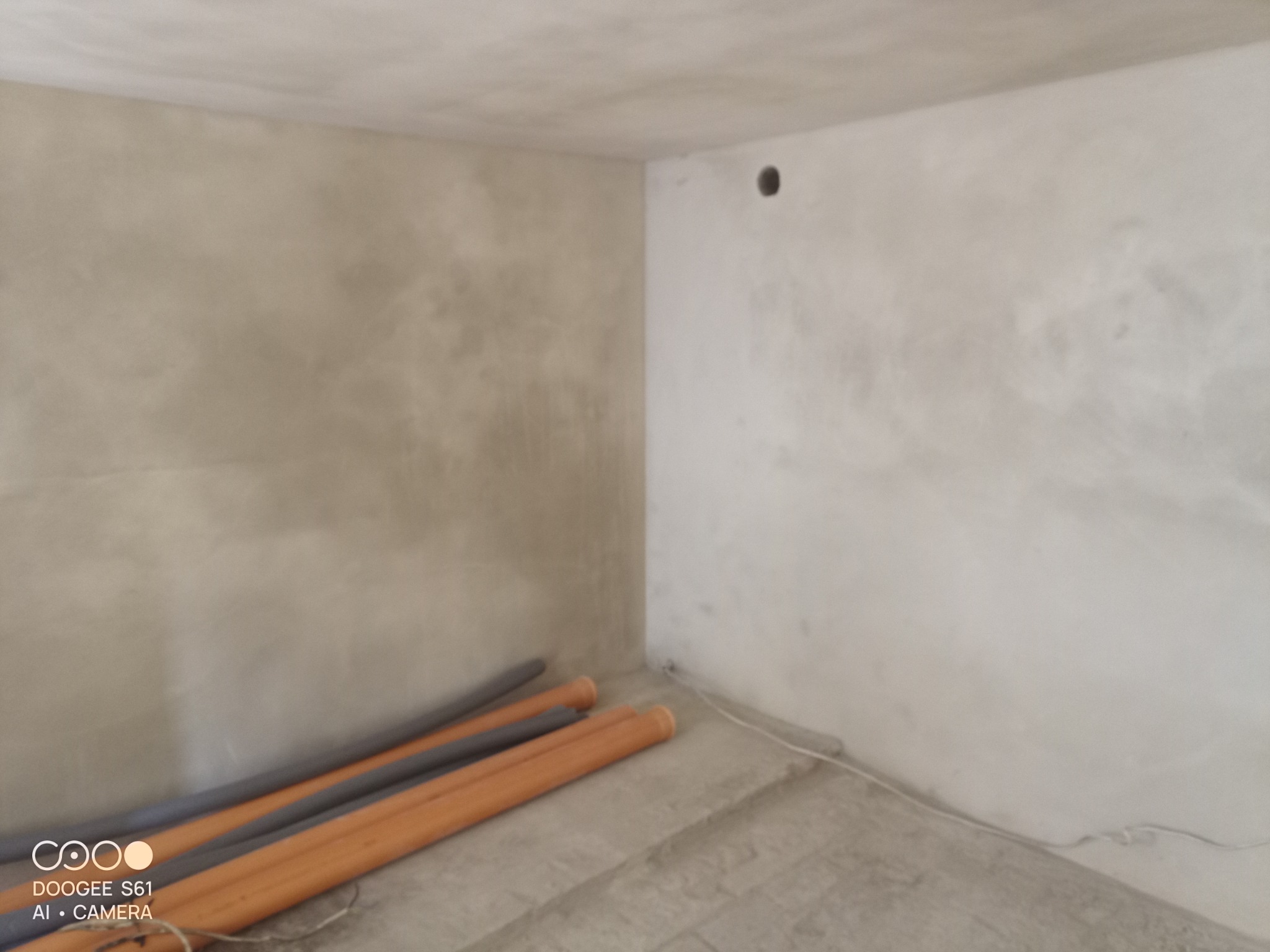 Świeżo otynkowane ściany w rogu pomieszczenia z widocznymi przewodami instalacyjnymi i rurami grzewczymi leżącymi na podłodze.