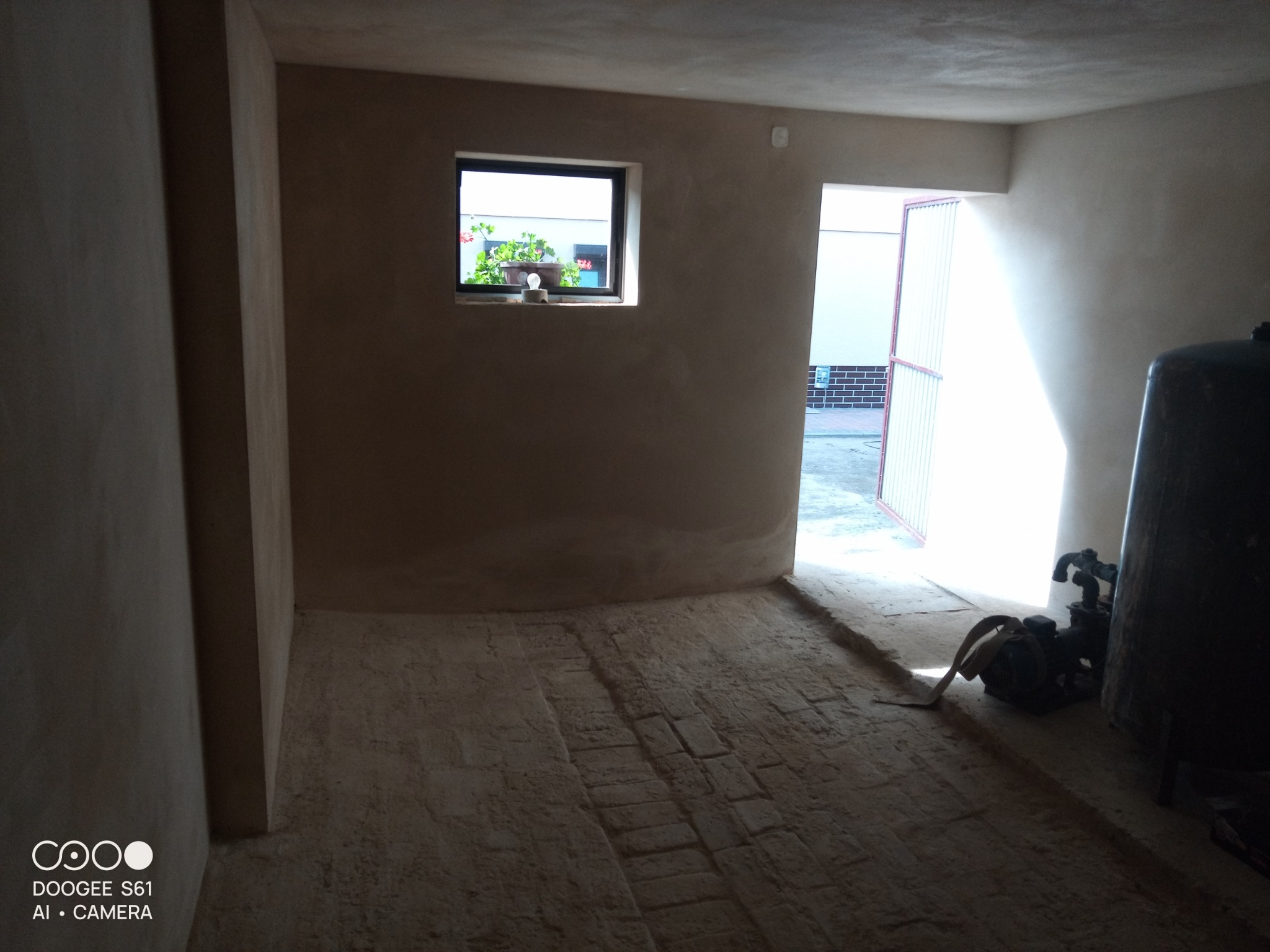 Wnętrze pomieszczenia po nałożeniu tynku, widoczna faktura ściany, okno z kwiatami i sprzęt do tynkowania w rogu.