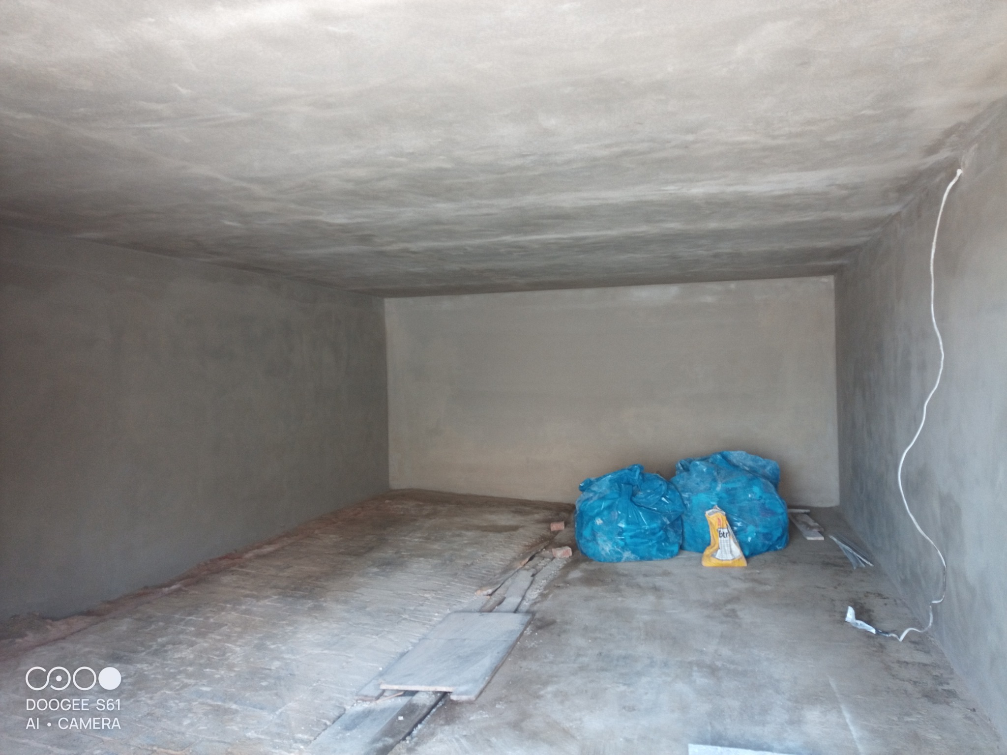 Wnętrze pomieszczenia po nałożeniu szarego tynku maszynowego na ściany i sufit, widoczne worki z odpadami budowlanymi i przewód elektryczny przy ścianie.