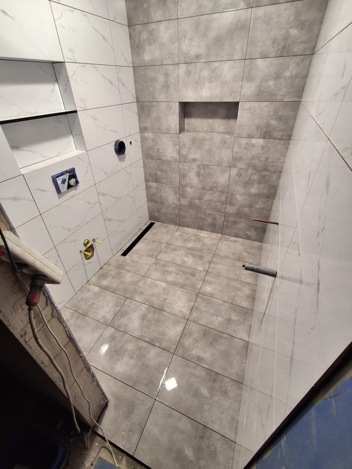 Wykończona łazienka z szarymi i białymi płytkami imitującymi marmur, z odpływem liniowym i wnęką w ścianie. Widoczne elementy instalacji.