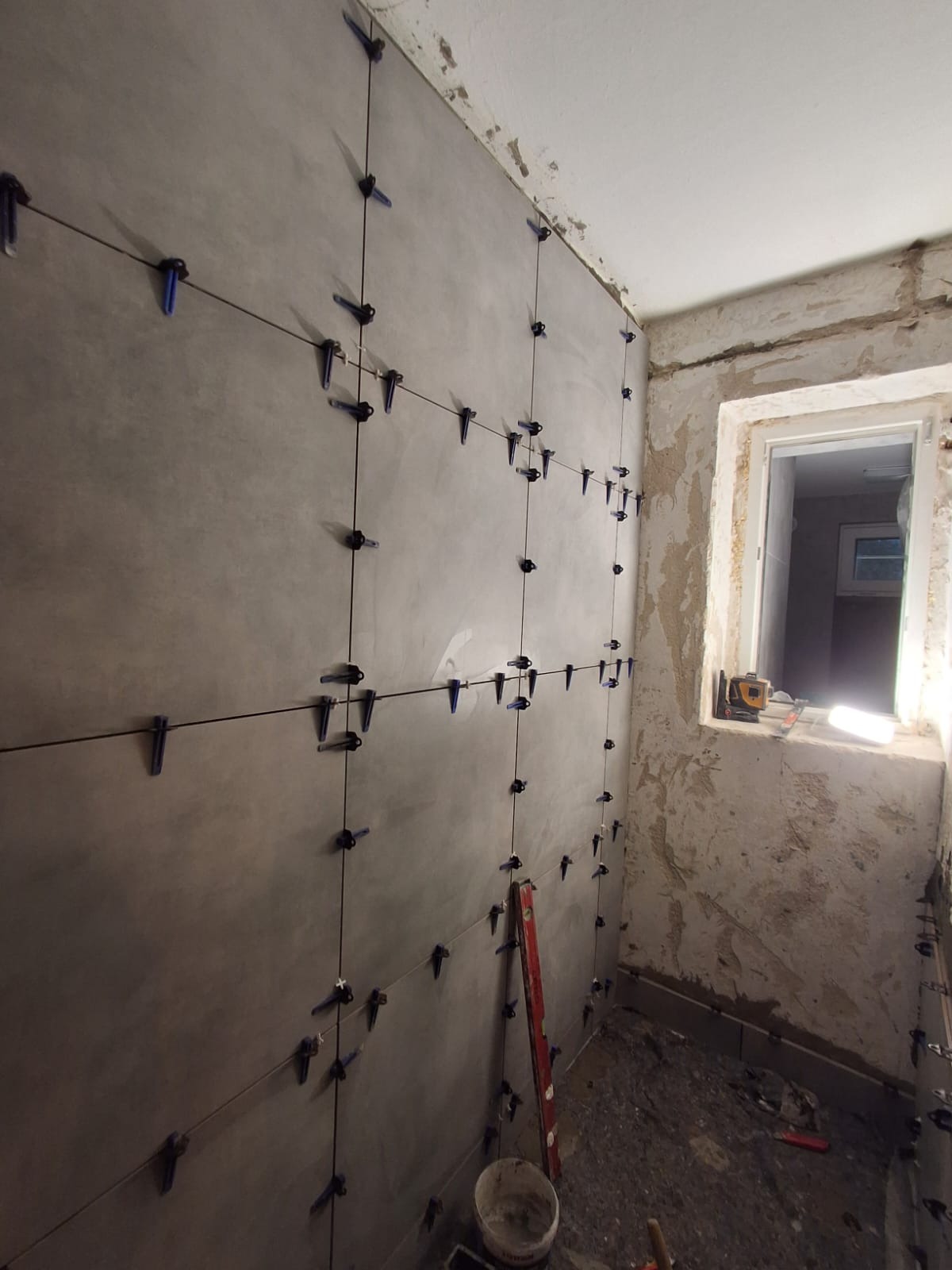 Ściana z szarych płytek w trakcie montażu, z użyciem systemu poziomowania płytek. Widoczne narzędzia i surowe wykończenie pomieszczenia.