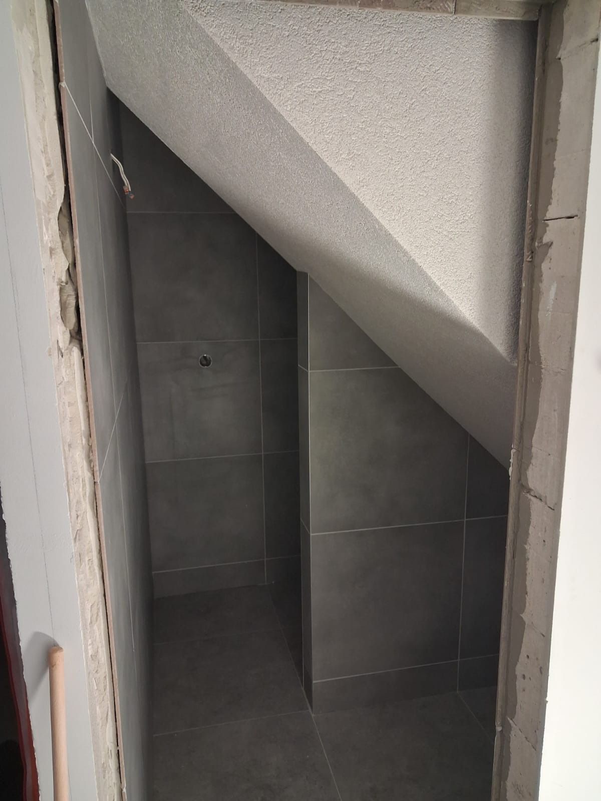 Wykończona łazienka na poddaszu z szarymi płytkami, widok z progu. Uwagę zwraca skośny sufit i precyzyjne ułożenie płytek w trudnej przestrzeni.