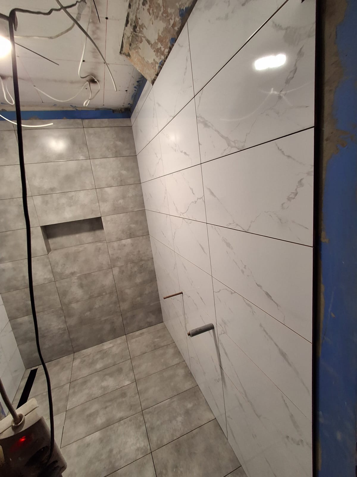 Wykończona łazienka z szarymi i białymi płytkami imitującymi marmur, widoczne wystające rury i niedokończony sufit. Ujęcie z dołu, podkreślające układ płytek.