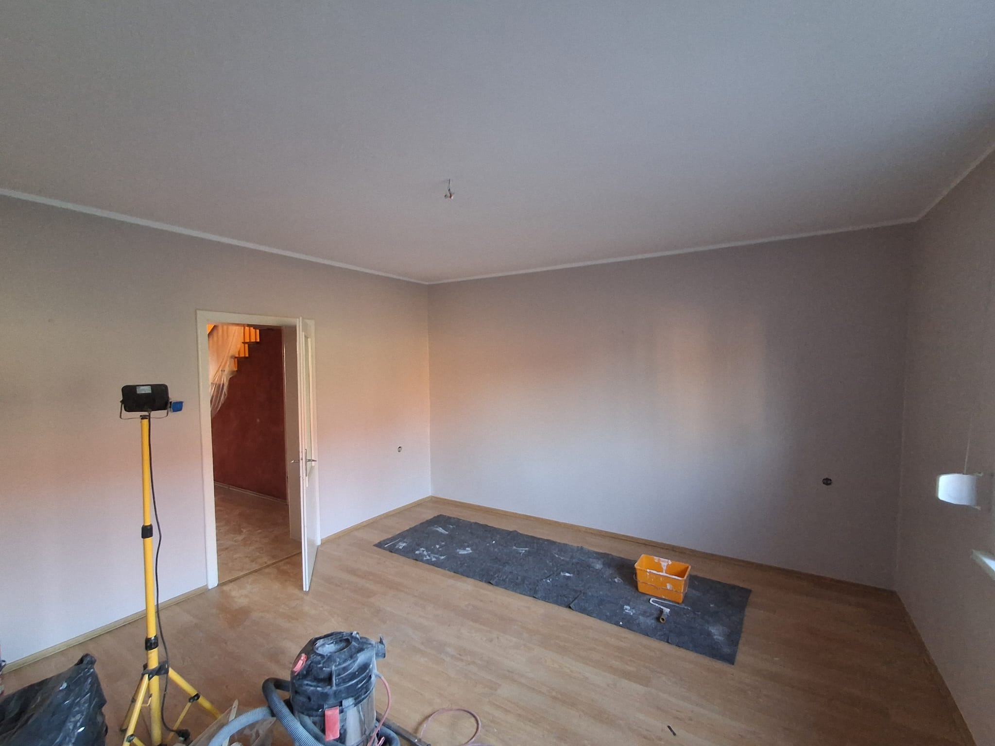 Pokój po malowaniu ścian na jasny odcień, widoczny sprzęt malarski, podłoga zabezpieczona, listwy przypodłogowe białe, w tle otwarte drzwi do innego pomieszczenia.