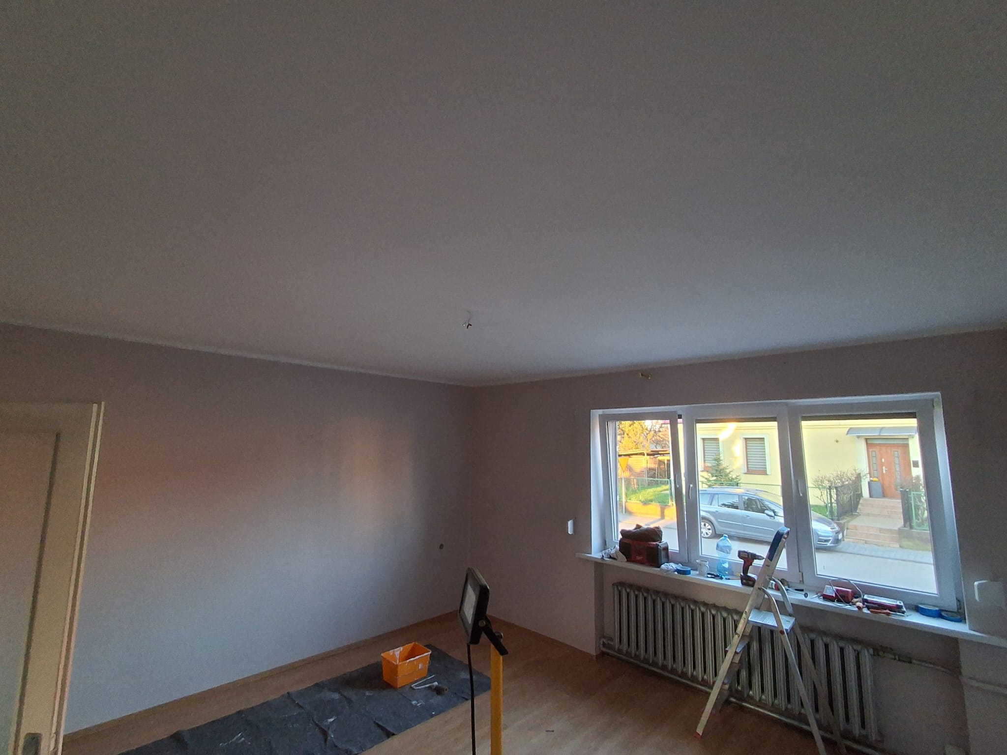 Pokój po malowaniu ścian na jasny odcień, widoczne narzędzia malarskie, drabina przy oknie, zabezpieczona podłoga. Świeżo pomalowane ściany i sufit.