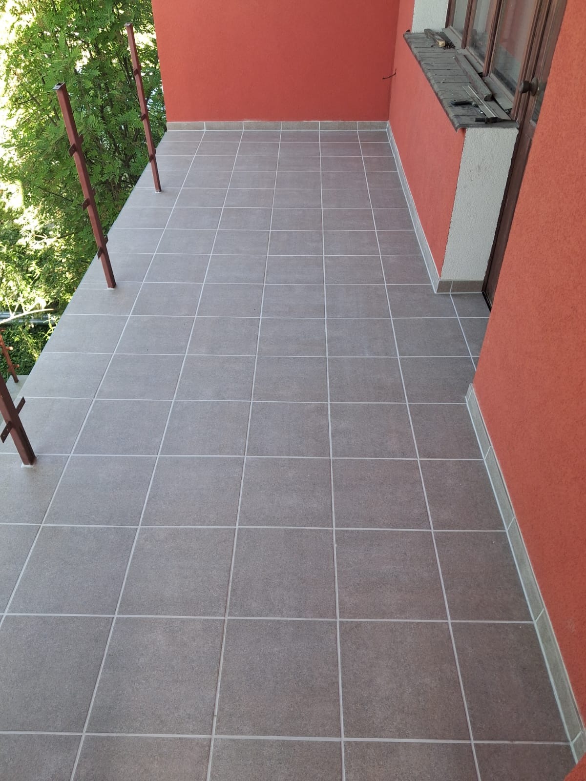 Wyłożony szarymi płytkami balkon z pomarańczową ścianą i metalową barierką. Widok na zieleń z zewnątrz. Płytki ułożone równo, z jasnymi fugami.