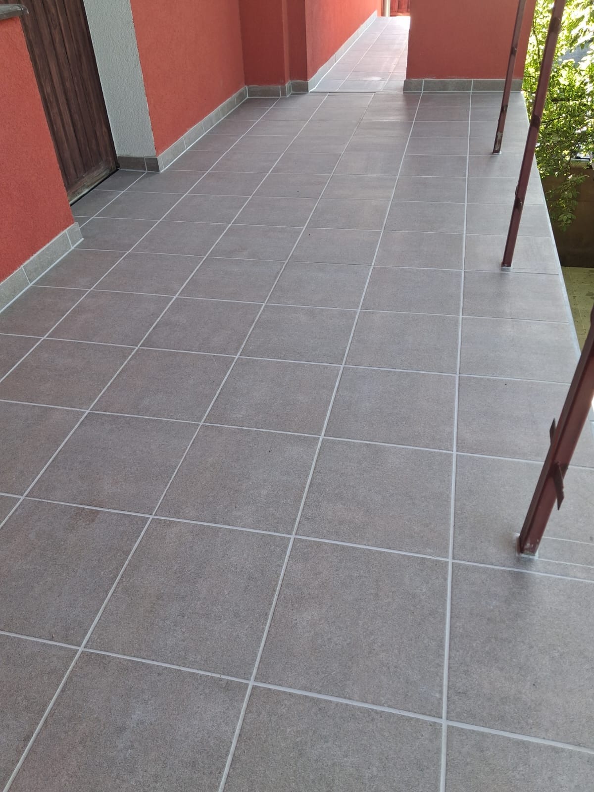 Ułożone, szare płytki podłogowe na zewnątrz budynku, z widocznymi brązowymi słupkami i fragmentem zieleni w tle. Widok z perspektywy.
