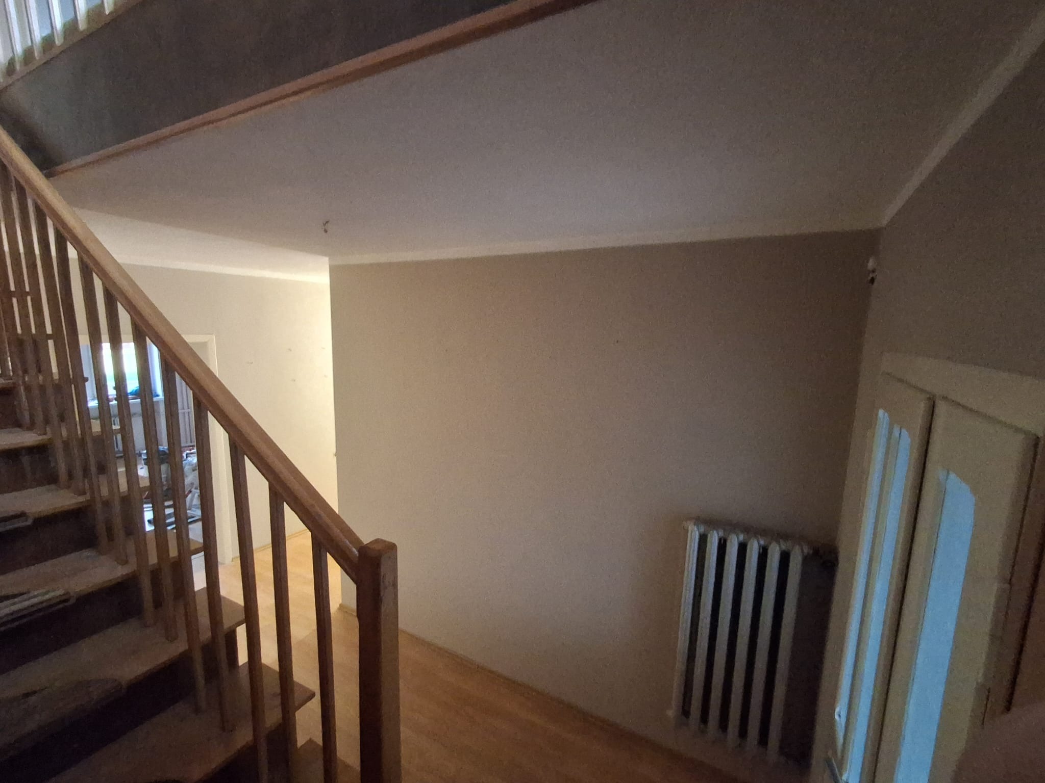 Jasne wnętrze domu w trakcie remontu: drewniane schody, pomalowane ściany w odcieniach beżu, grzejnik żeberkowy i częściowo widoczne drzwi.