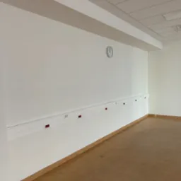 Wnętrze wykończonego pomieszczenia z białymi ścianami, brązową podłogą, listwą przypodłogową i zegarem na ścianie. Widoczne gniazdka elektryczne w listwie. Sufit podwieszany.