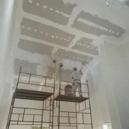 Pracownik szlifuje połączenia płyt na suficie podwieszanym z płyty gipsowo-kartonowej, stojąc na rusztowaniu w wysokim pomieszczeniu z oknem.