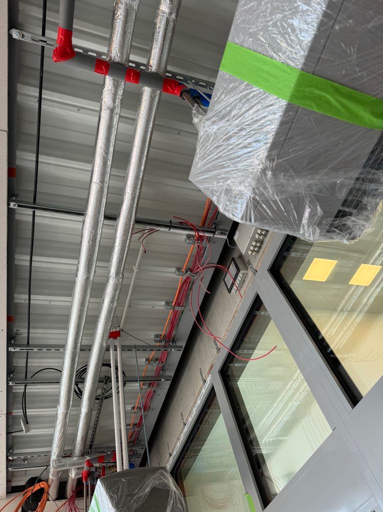 Widok z dołu na instalacje pod sufitem: rury owinięte folią aluminiową z czerwonymi złączkami, kable elektryczne w pomarańczowej izolacji, szyny montażowe oraz okno z widocznymi żółtymi lampami...