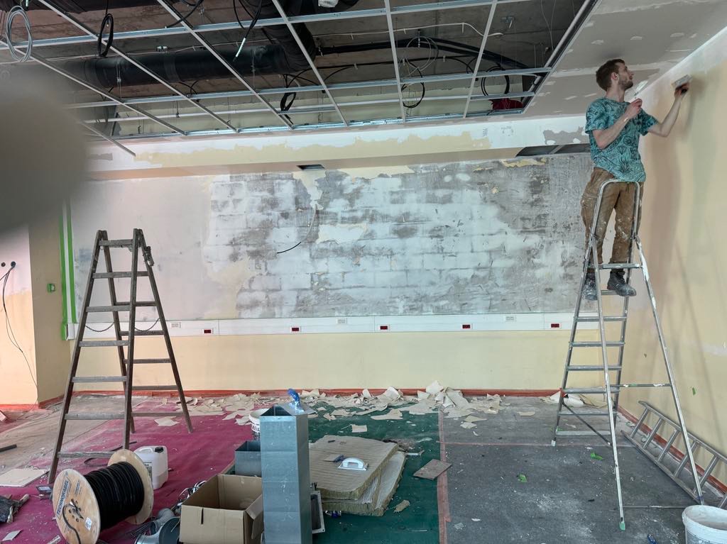Mężczyzna na aluminiowej drabinie nanosi gładź na ścianę w remontowanym pomieszczeniu z odsłoniętą konstrukcją sufitu, widoczne narzędzia i materiały budowlane na podłodze.
