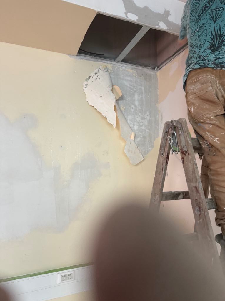 Przygotowanie ściany do gładzi: usunięte fragmenty starej farby i tapety, widoczny fragment sufitu podwieszanego, pracownik na drewnianej drabinie podczas remontu.