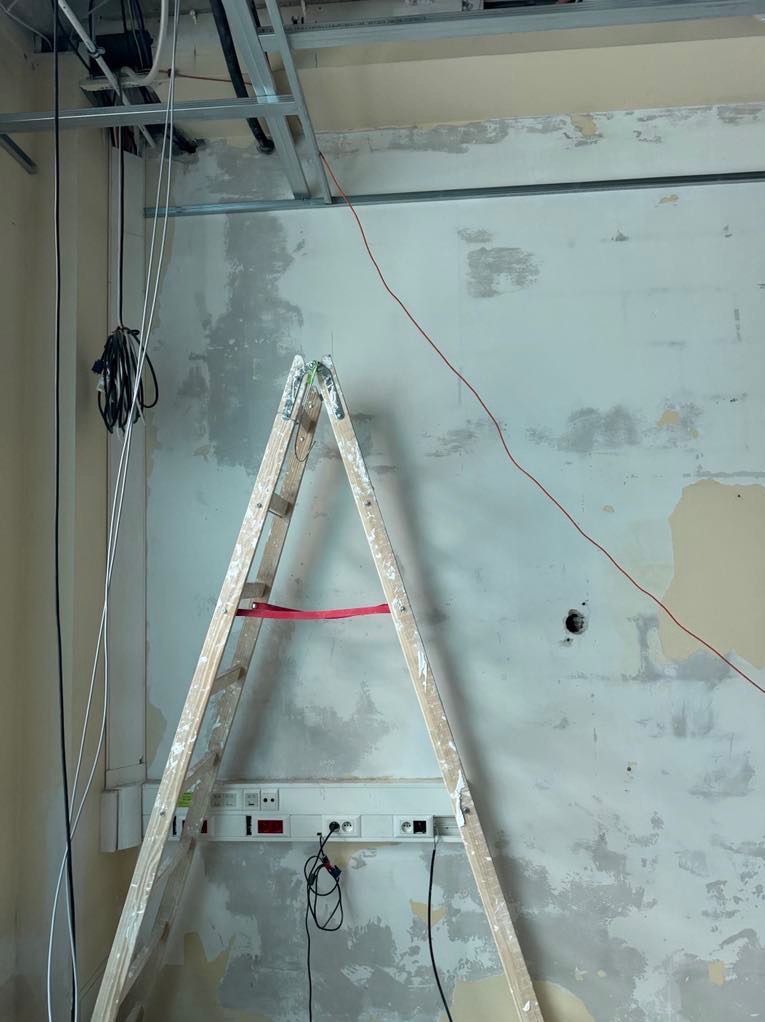 Ściana w trakcie prac przygotowawczych z widocznymi śladami po poprzednich warstwach, drabina malarska oparta o ścianę, instalacja elektryczna w korycie i metalowa konstrukcja podwieszanego sufitu.